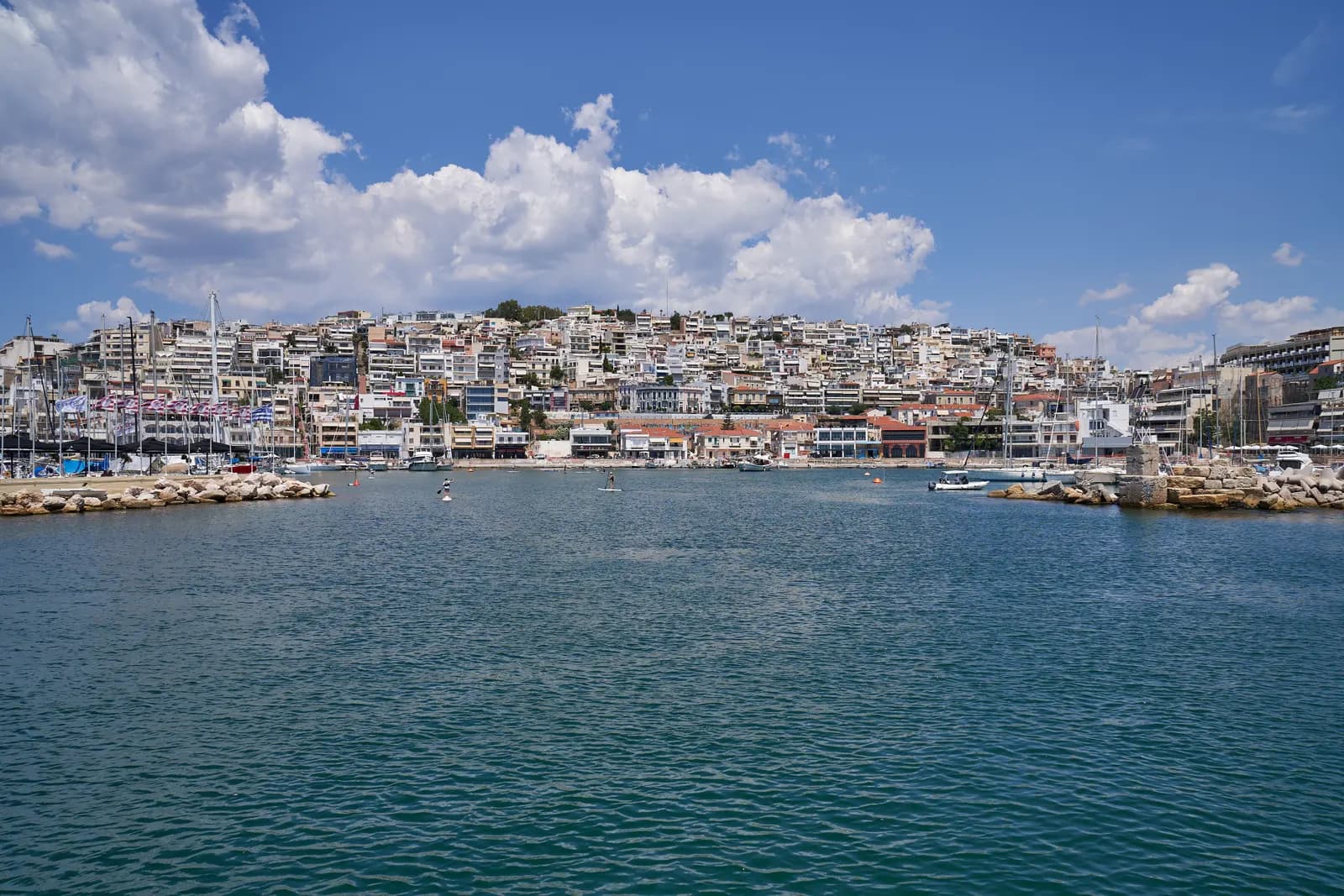 The entrance to Mikrolimano harbour in Piraeus, with moored yachts and white apartment buildings climbing Kastella hill under a summer sky