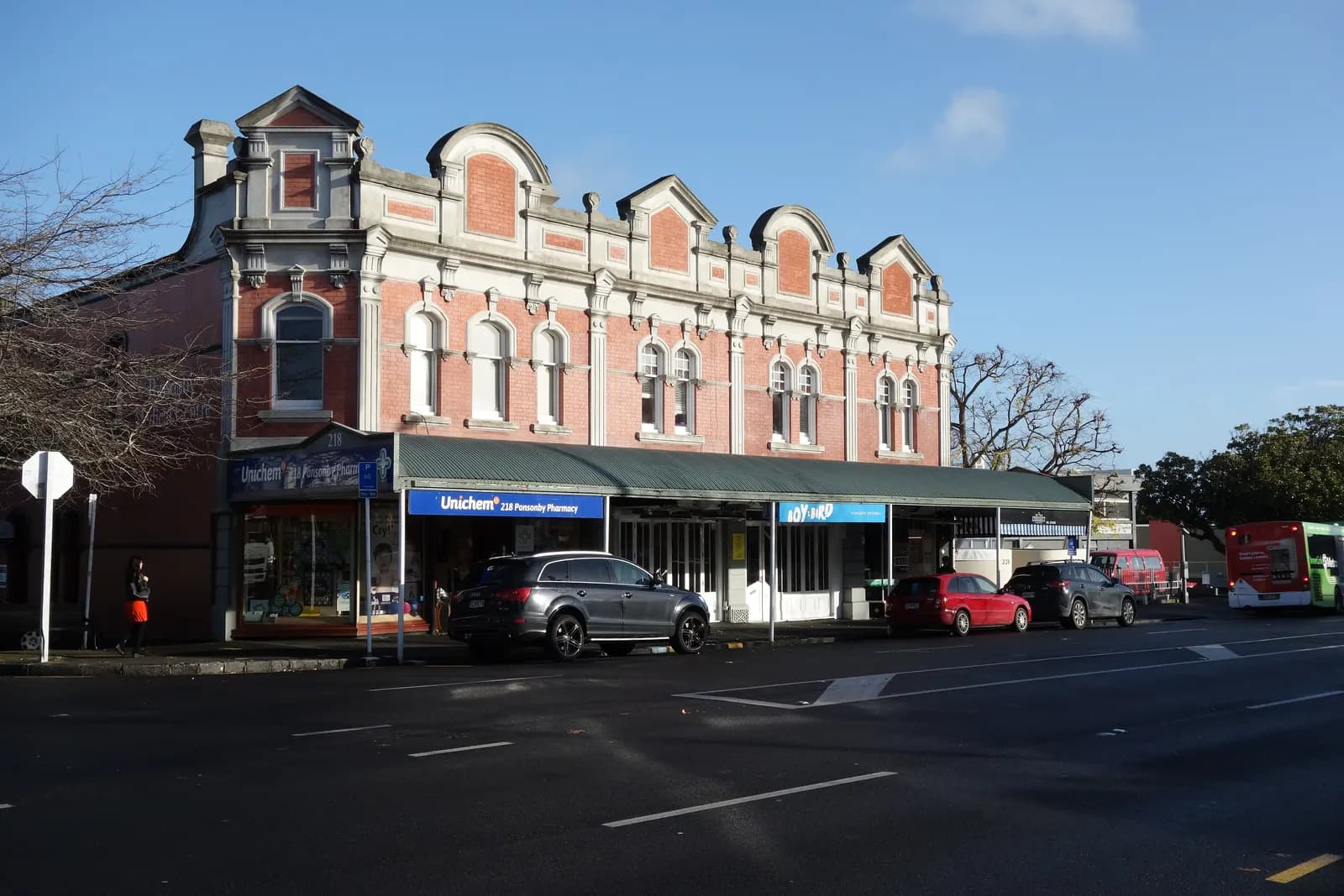 A two-storey brick building with shops at street level on Ponsonby Road, with cars parked in front and a bus further along