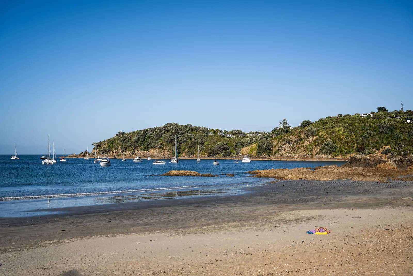 The sandy beach of Oneroa Bay on Waiheke Island, with boats at anchor and a bush-covered headland with houses