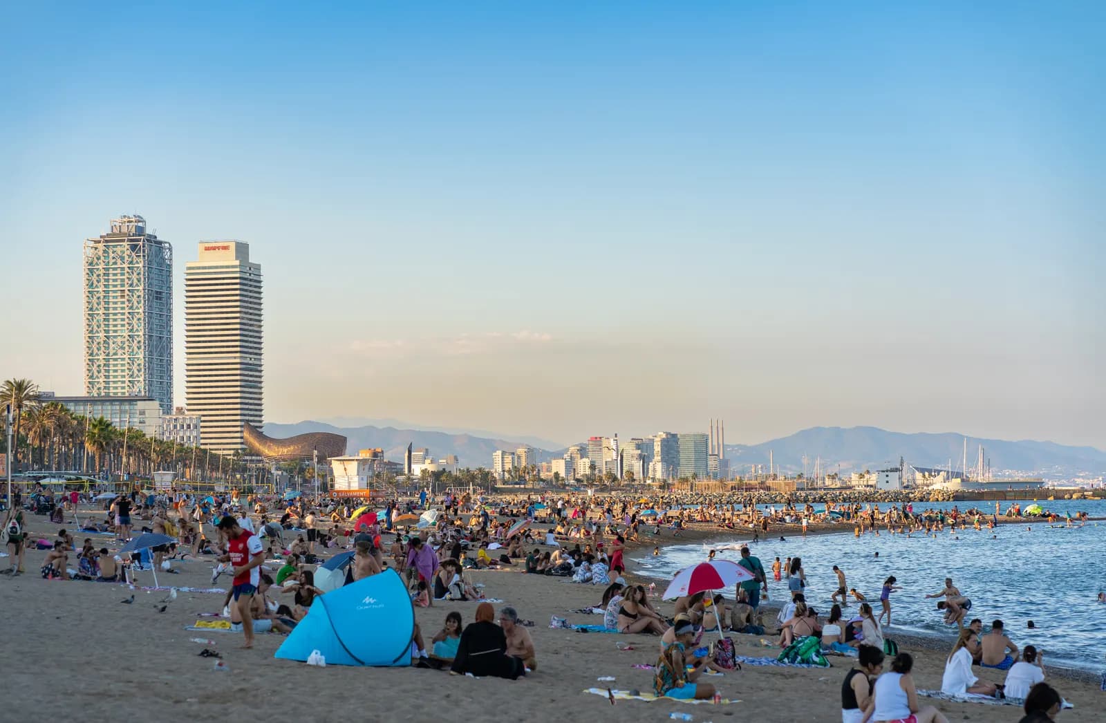 A busy summer afternoon on Barceloneta beach, with the two Olympic Port towers and the mountains behind.