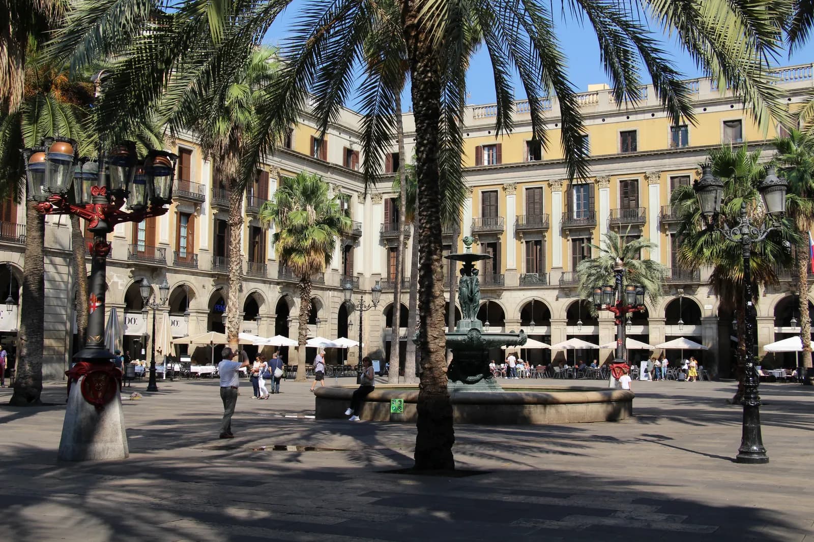 Plaça Reial in the Gothic Quarter with palm trees, the central fountain, arcades and café terraces in sunshine.