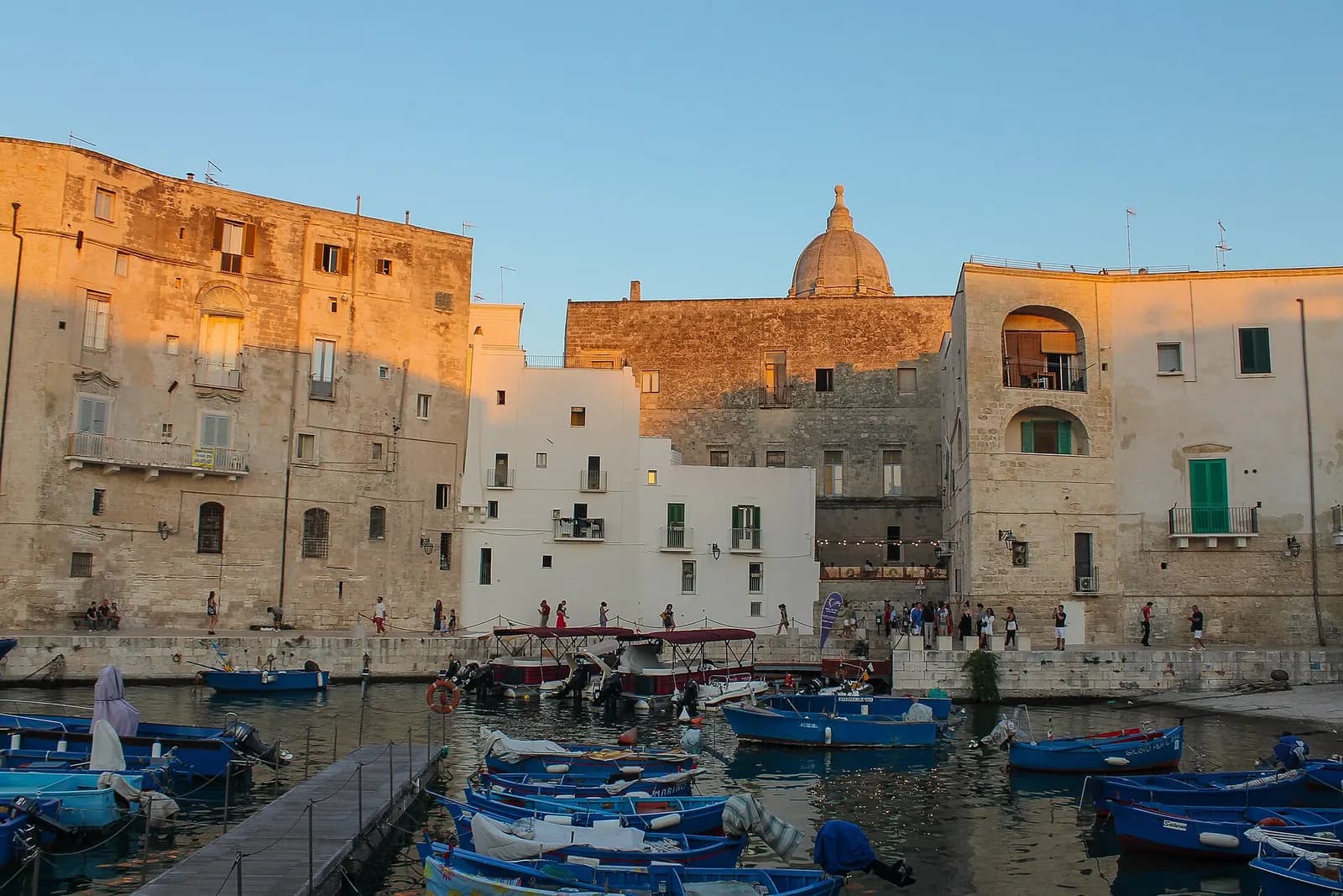 Blue fishing boats in the old harbour of Monopoli below stone houses and the dome of the church of Santa Teresa in evening light