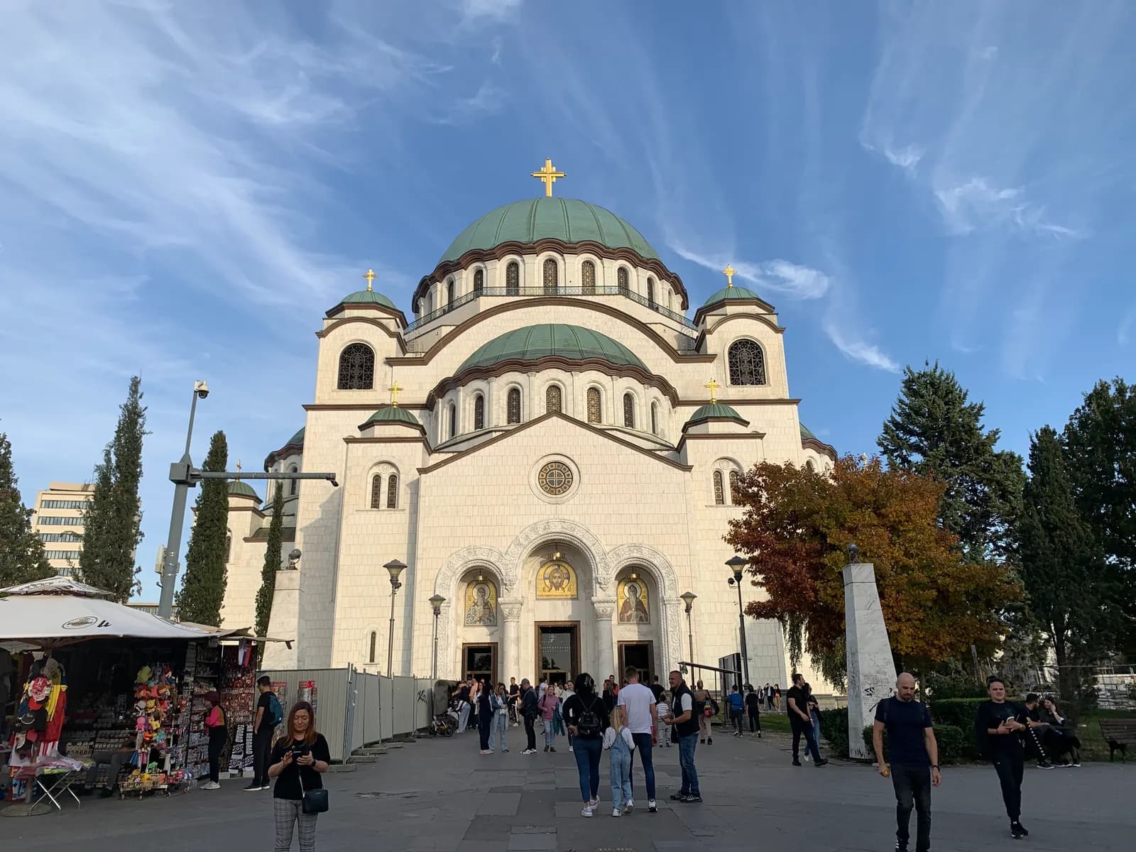 The white Church of Saint Sava on Vračar in Belgrade, with green domes and gold crosses, visitors on the square in front