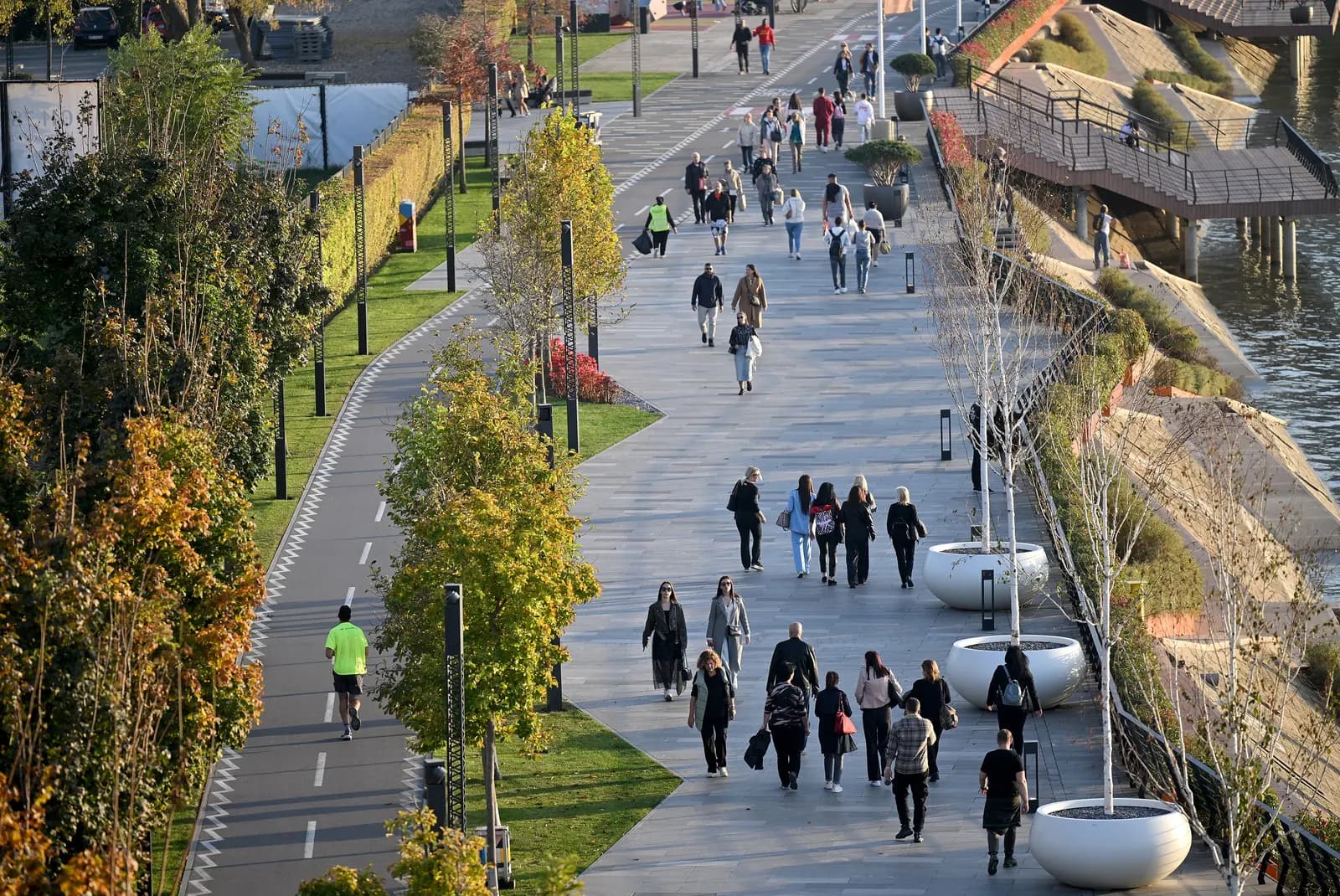 People walking on the Sava Promenade in Belgrade, a wide riverside path with a cycle lane, young trees and planters by the water