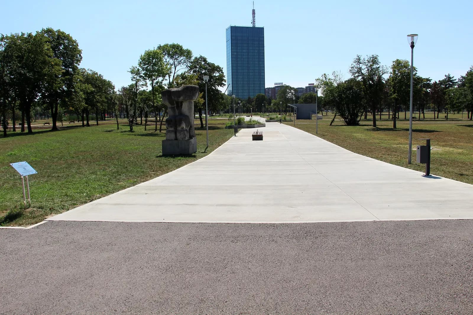 A paved path through Ušće park in New Belgrade, with a stone sculpture on the lawn and the glass Ušće Tower behind the trees