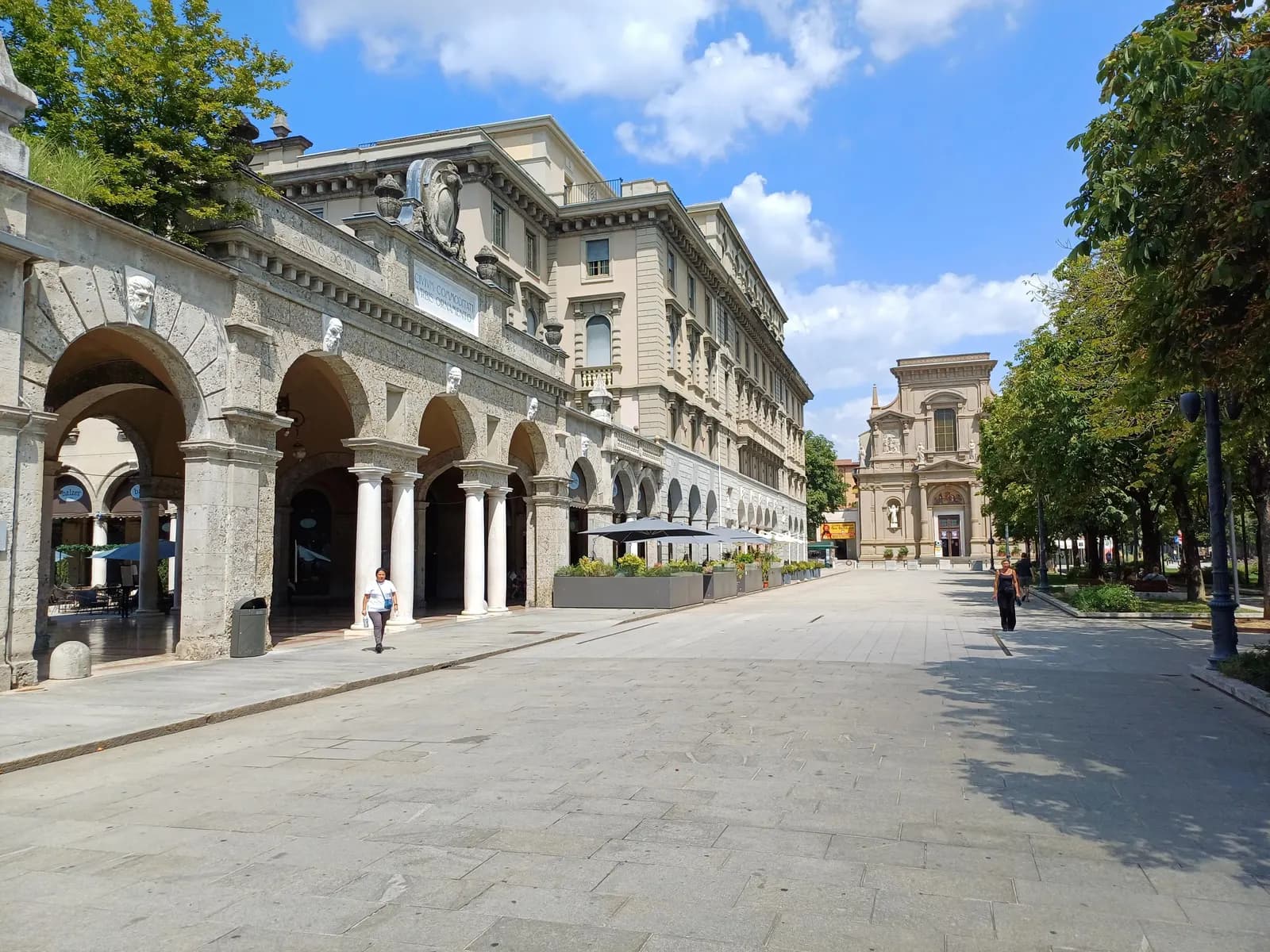 The Sentierone in the lower town, a paved promenade with stone arcades on one side and trees on the other, and the church of San Bartolomeo at the far end.