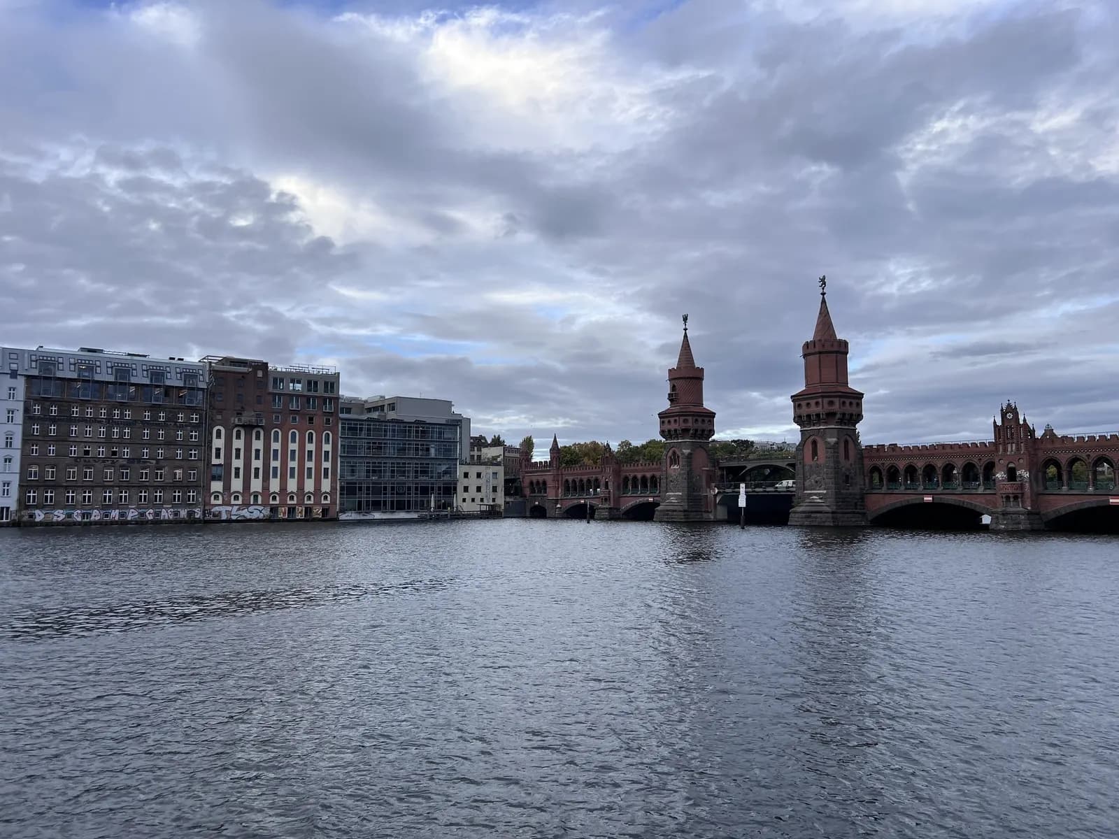 The red-brick Oberbaum Bridge over the Spree seen from the east, with warehouse buildings on the Friedrichshain bank under a cloudy sky