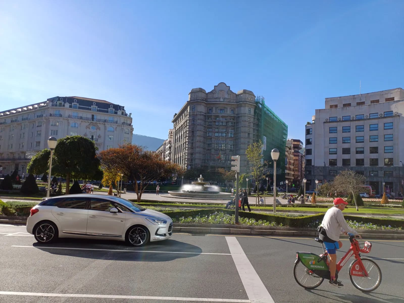 Plaza Moyua in Abando, with a car and a cyclist on the road in front of its gardens, a fountain and large stone buildings.