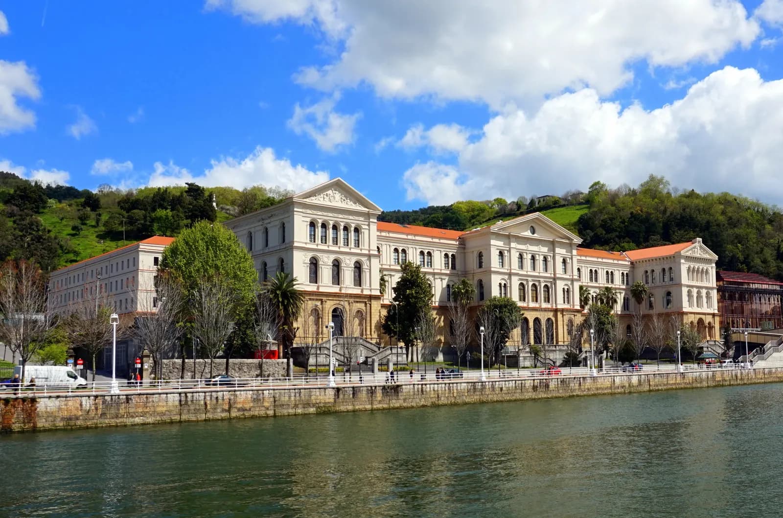 The pale stone main building of the University of Deusto on the estuary bank, with green hills behind.