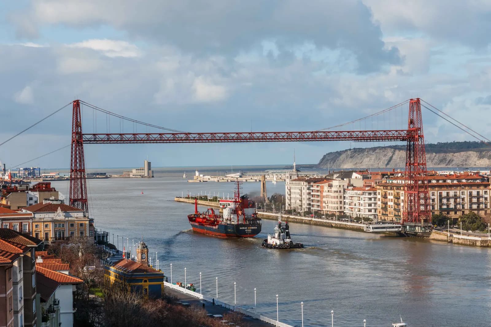 The red iron Bizkaia transporter bridge spanning the estuary mouth between Getxo and Portugalete, with a small cargo ship passing below.