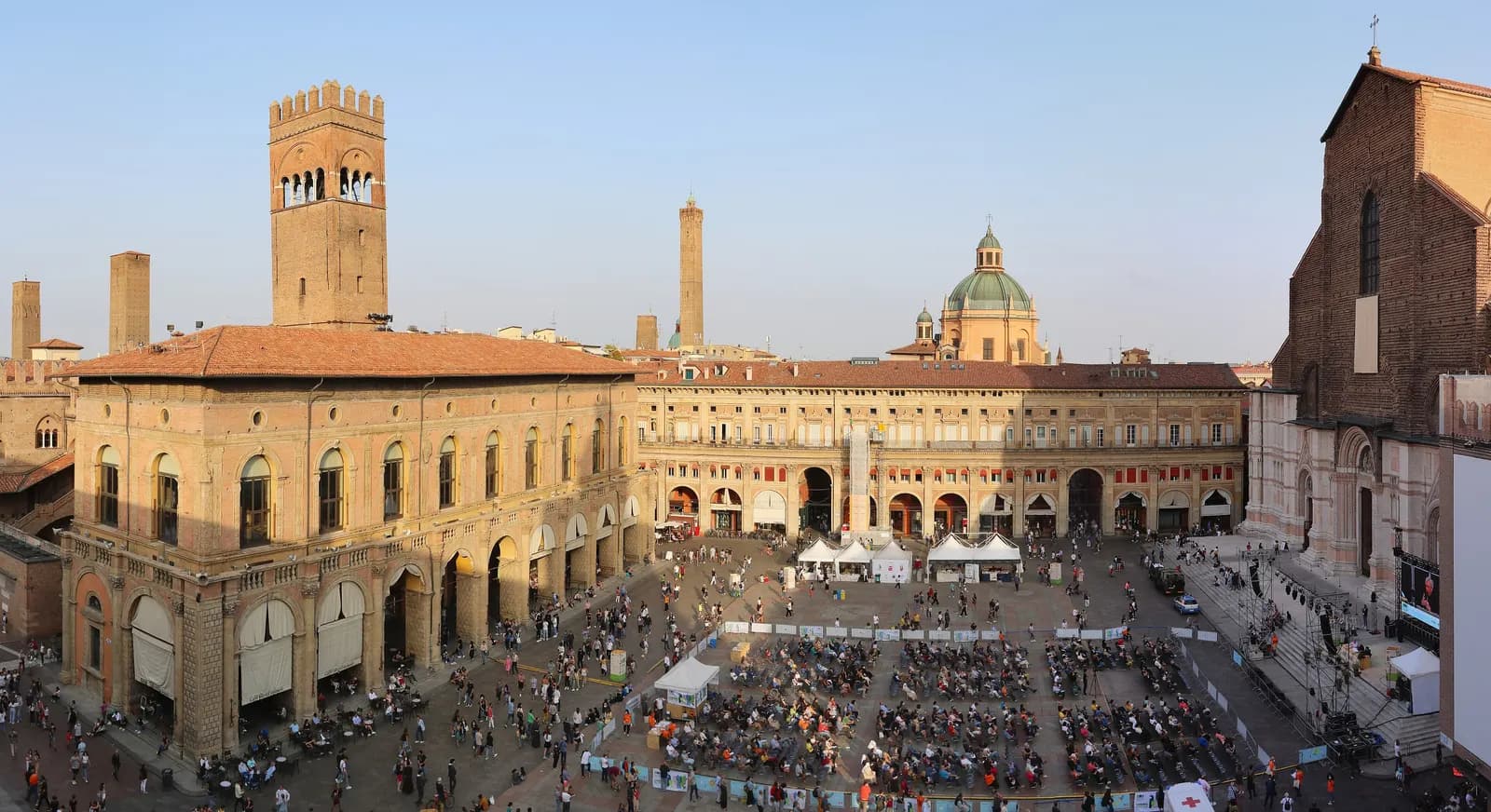 Piazza Maggiore seen from above in late afternoon: the Palazzo del Podesta and its tower, the arcaded Palazzo dei Banchi, the flank of San Petronio and rows of chairs set out for an open-air screening.