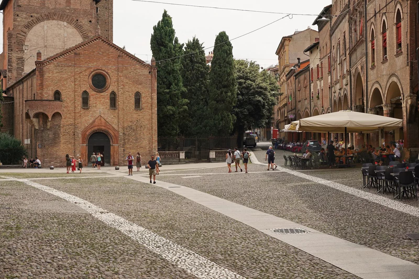 The cobbled triangle of Piazza Santo Stefano in Bologna, with the brick church front on the left, arcaded houses on the right and cafe tables under a large umbrella.