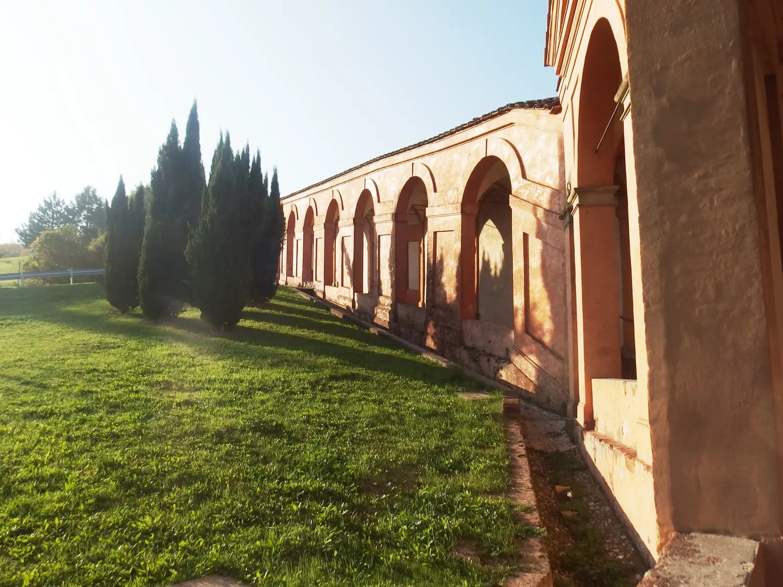 A stretch of the arcaded Portico di San Luca climbing the hill above Bologna in low sun, with cypresses and grass beside it.