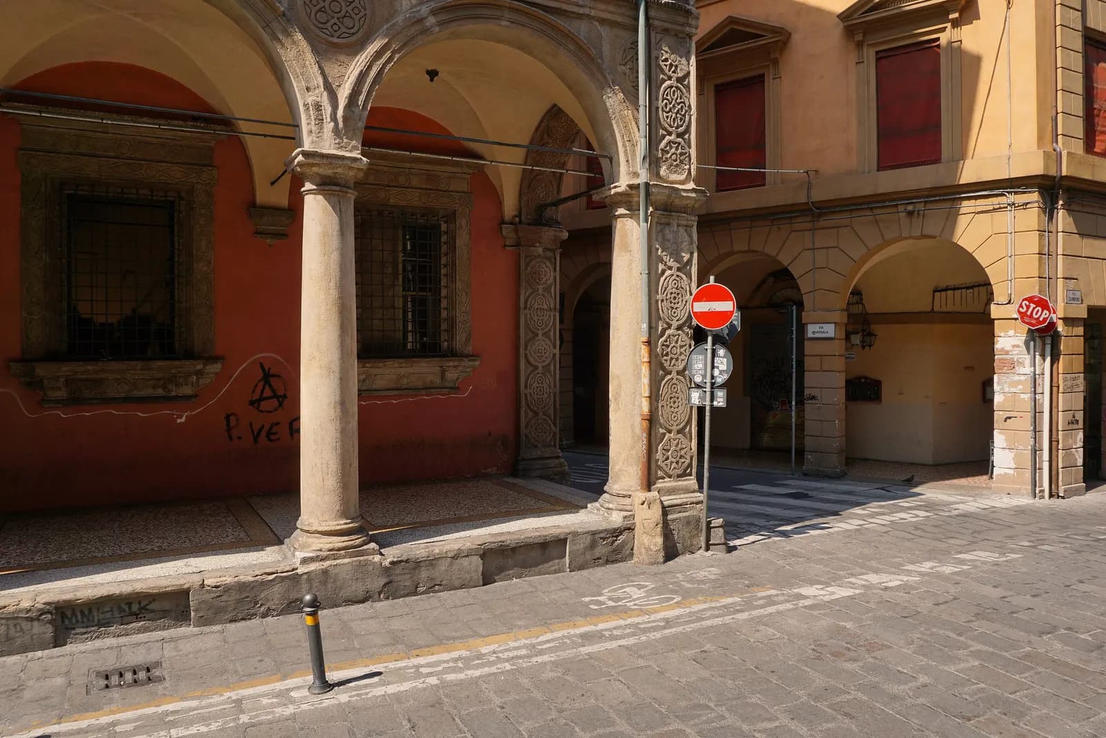 A portico of carved stone columns on Via Zamboni in Bologna at the corner of Via Marsala, with a no-entry sign, a stop sign and an empty cobbled street.