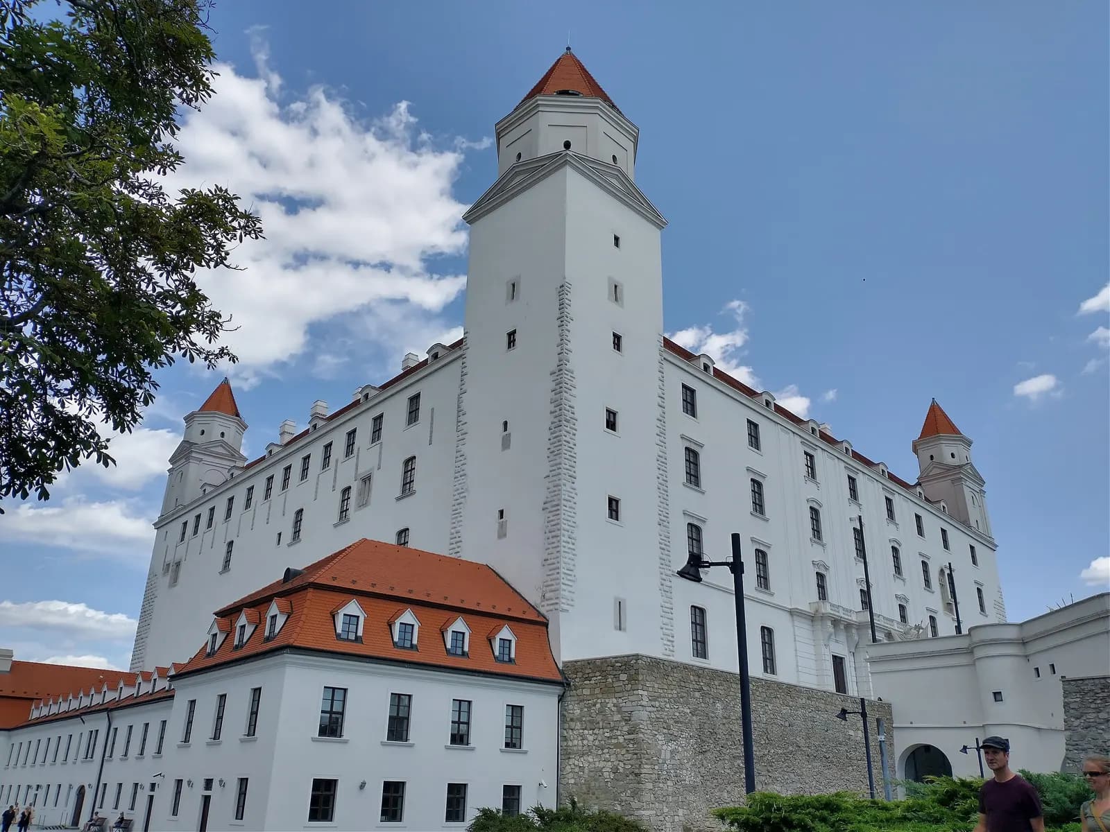 The white walls and corner towers of Bratislava Castle with red roofs, seen from below on the castle grounds under a blue sky