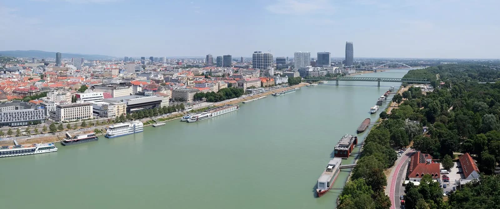The Danube in Bratislava seen from above, with river cruise boats moored along the Old Town embankment on the left, high-rise towers behind and the wooded Petržalka bank on the right