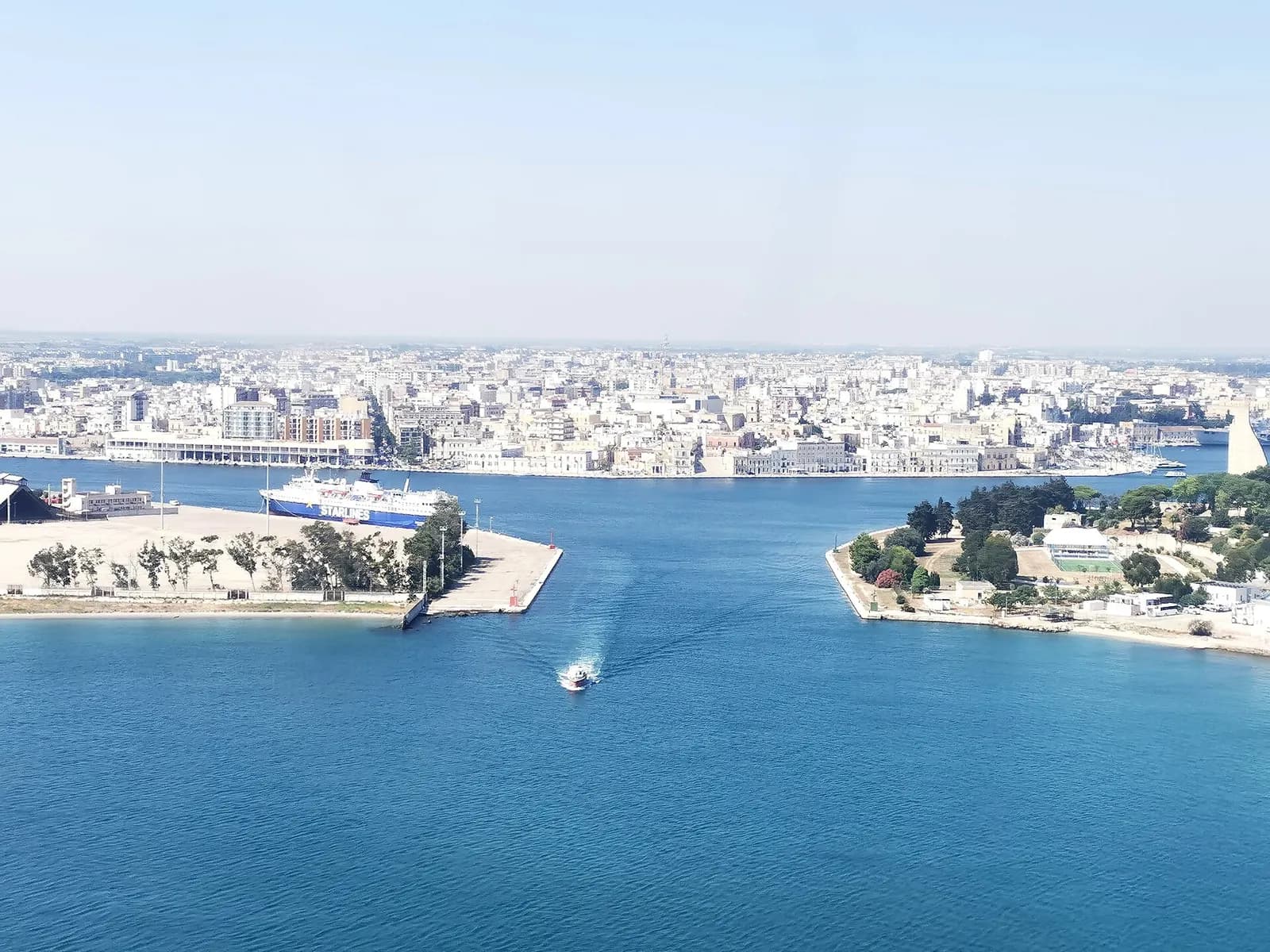 The entrance to Brindisi harbour seen from above, with a ferry at the quay, a small boat heading in and the city beyond