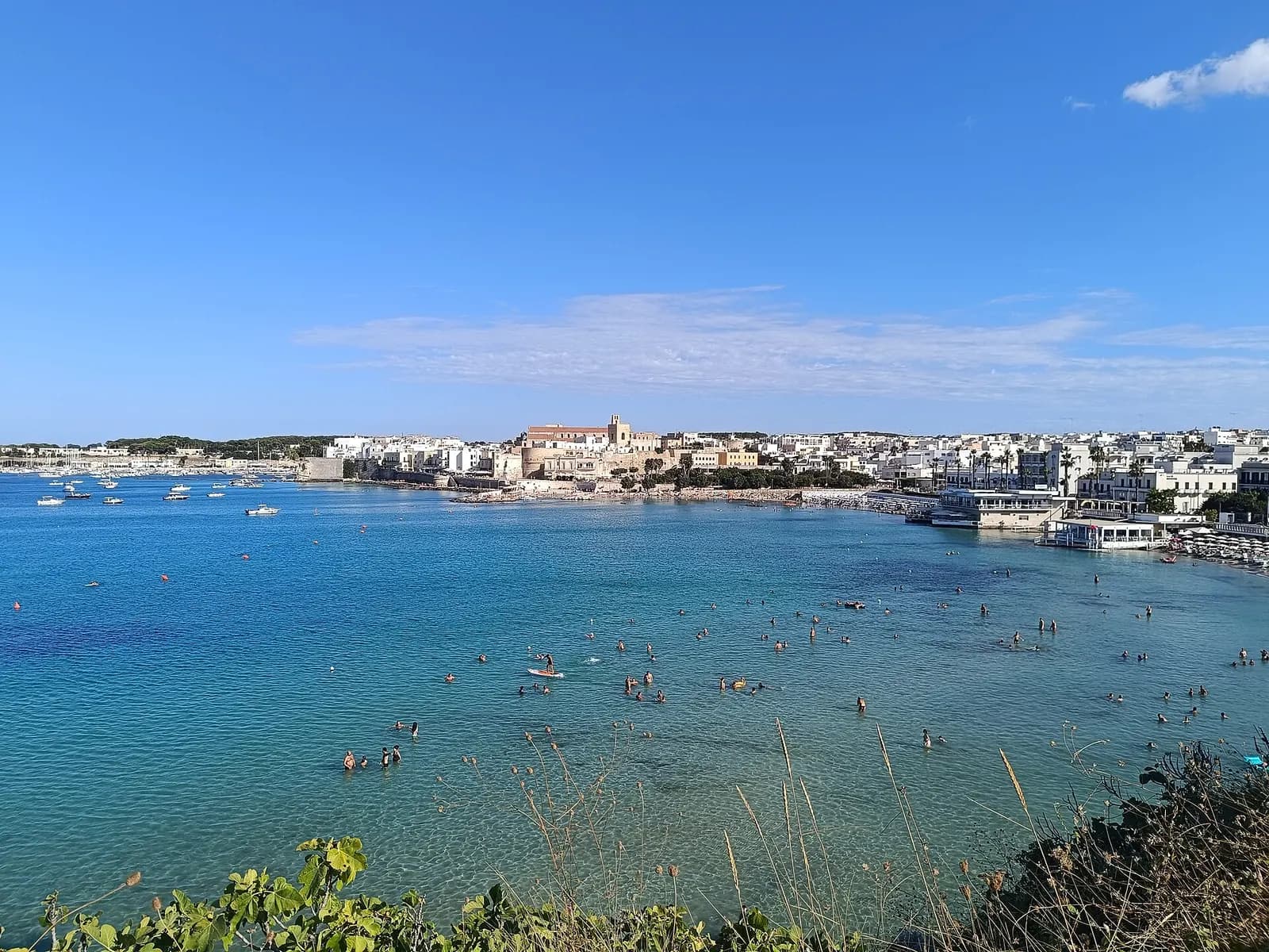 The bay of Otranto seen from above, with swimmers in clear shallow water and the old town and harbour along the far shore