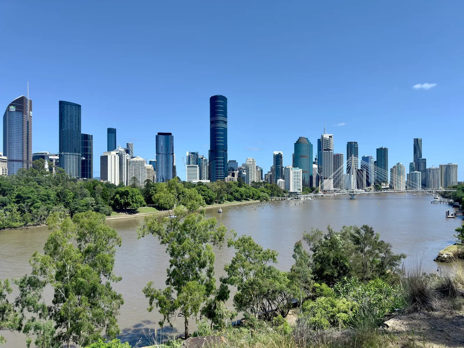 The Brisbane CBD skyline across the Brisbane River, seen from Kangaroo Point, with trees on both banks and the Kangaroo Point Bridge on the right