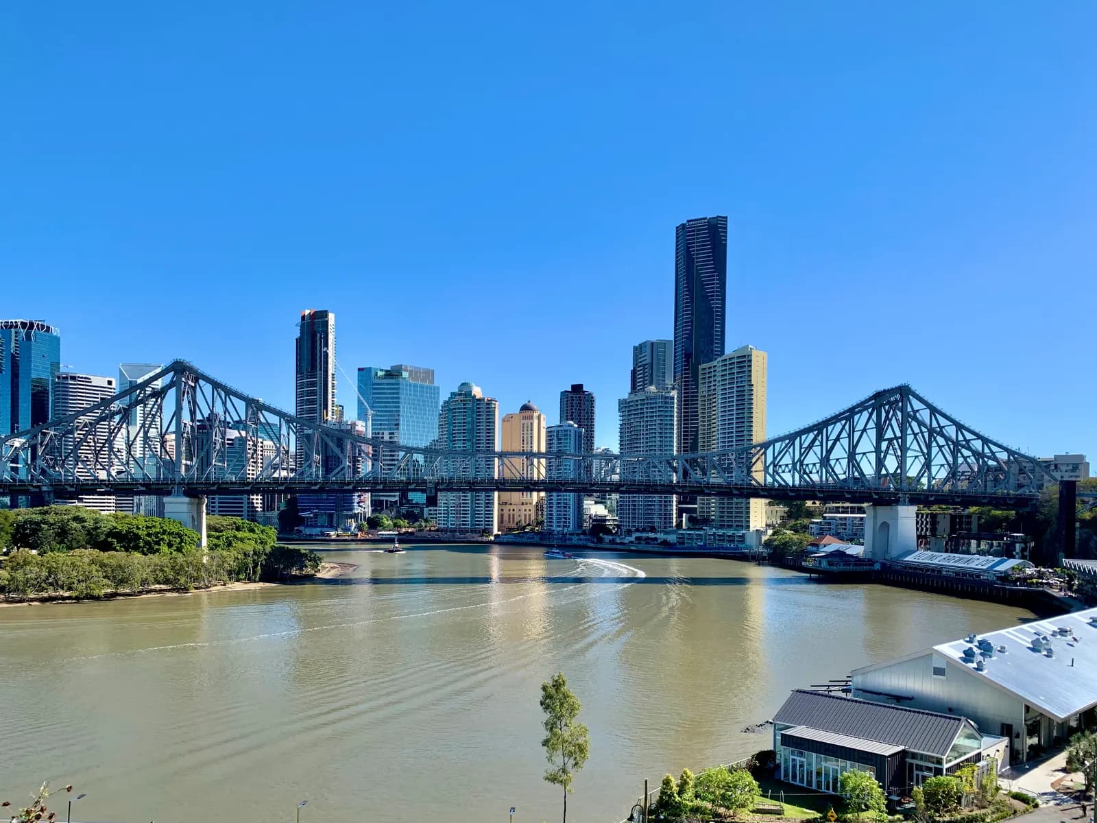 The Story Bridge over the Brisbane River, with the towers of the Brisbane CBD behind it and riverside buildings in the foreground, under a clear blue sky