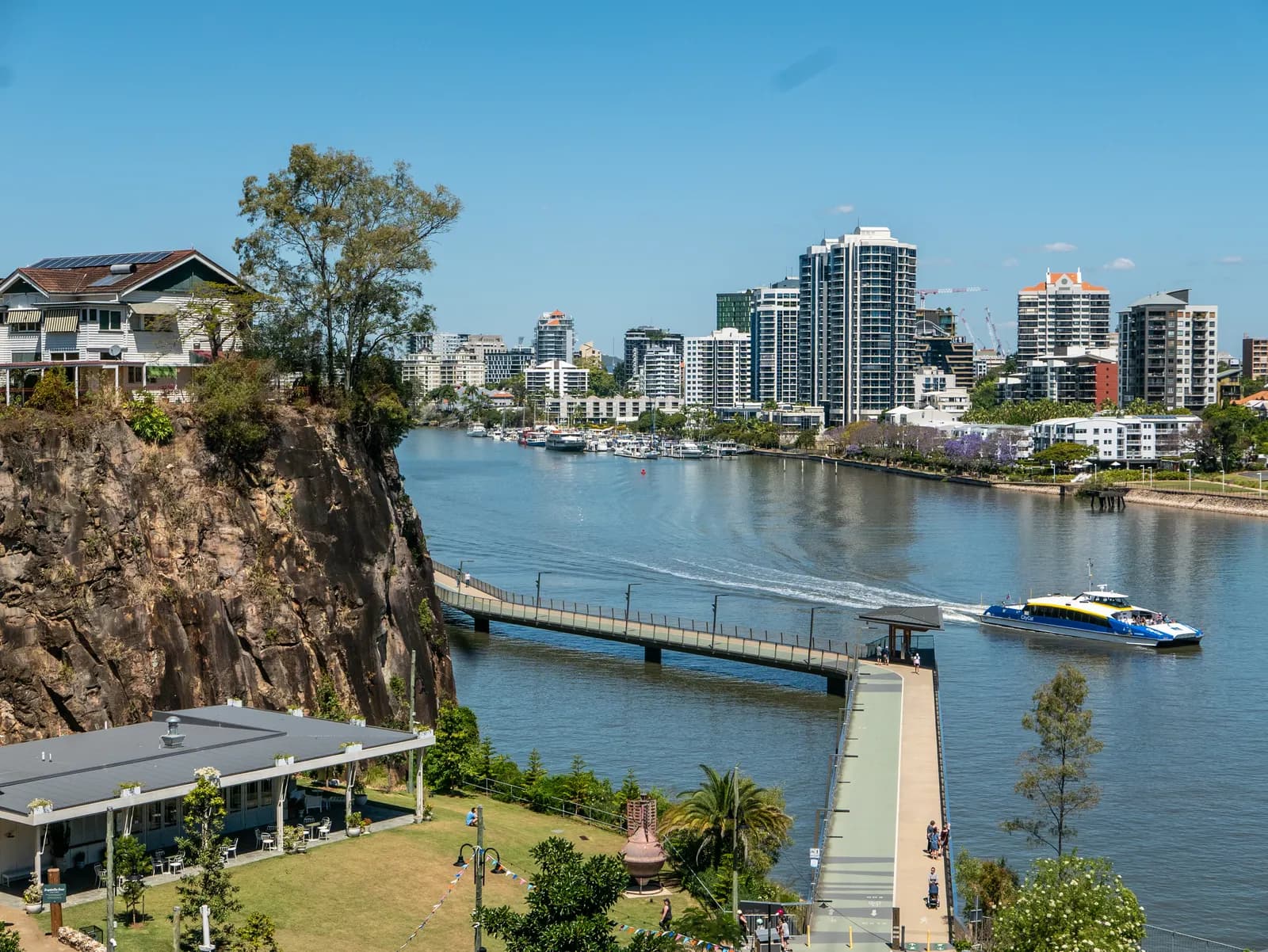 The New Farm Riverwalk running along the Brisbane River below a cliff, with a CityCat ferry, moored boats and apartment towers along the riverbank