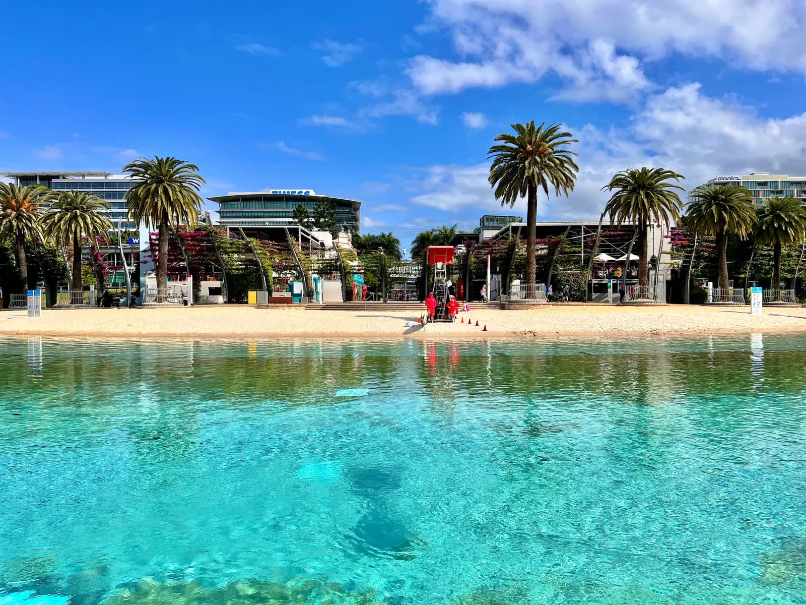 Streets Beach at South Bank Parklands, a lagoon of clear water with a sandy beach, palm trees, a red lifeguard tower and buildings behind