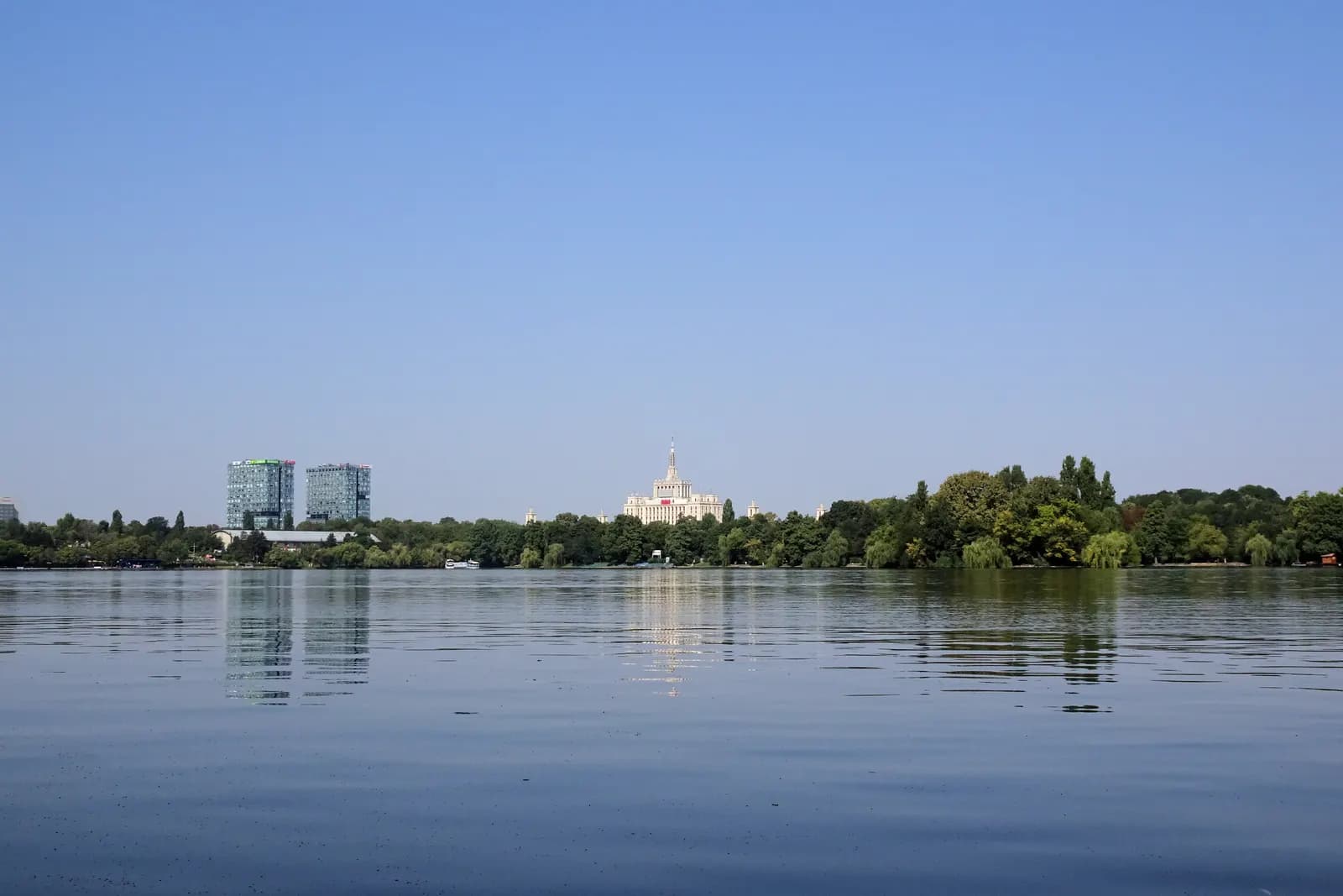 The calm water of Lake Herăstrău in north Bucharest, with a line of trees on the far shore, a white tower building and two glass office blocks behind