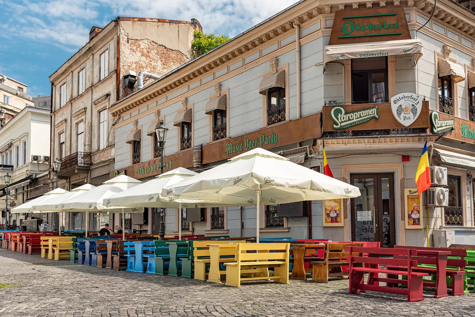 Empty café terraces with white umbrellas and colourful wooden benches on a cobbled street in Bucharest's Old Town, in front of a pale 19th-century corner building