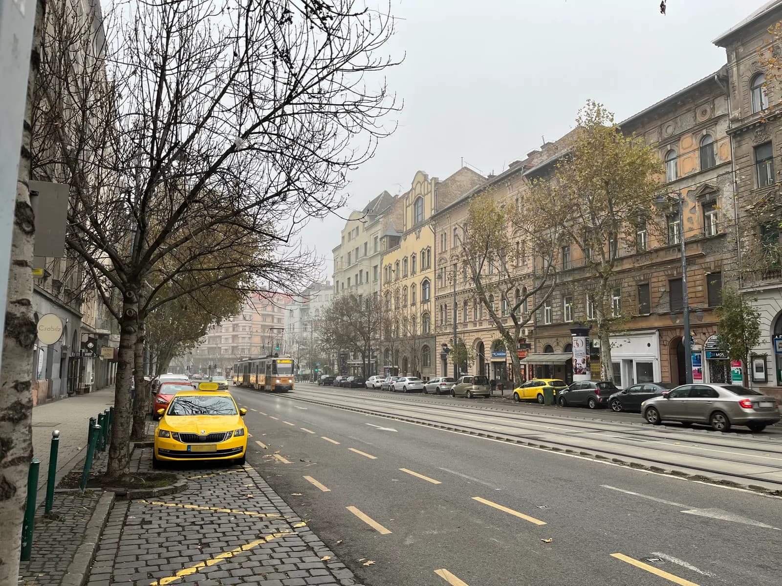 Bartók Béla út in Újbuda on an overcast day, with tram tracks, a tram in the distance, cars parked along the kerb and apartment buildings