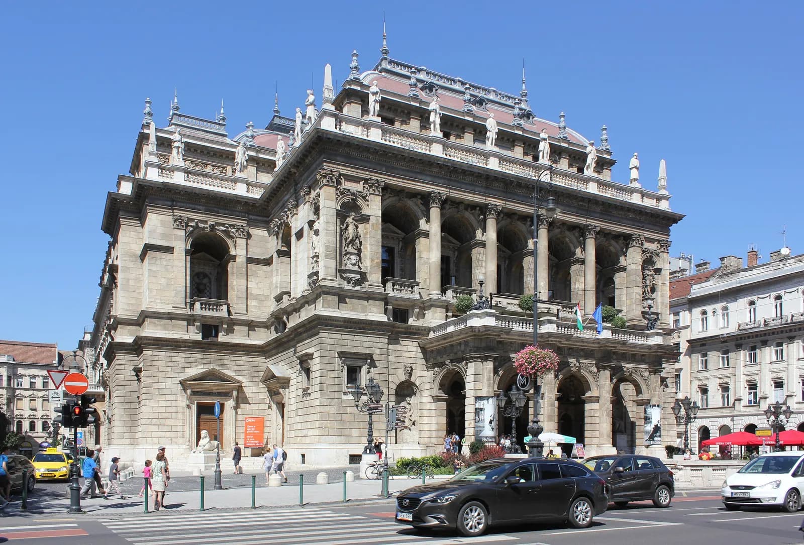 The neo-Renaissance Hungarian State Opera House on Andrássy út, with cars passing on the avenue in front under a blue sky