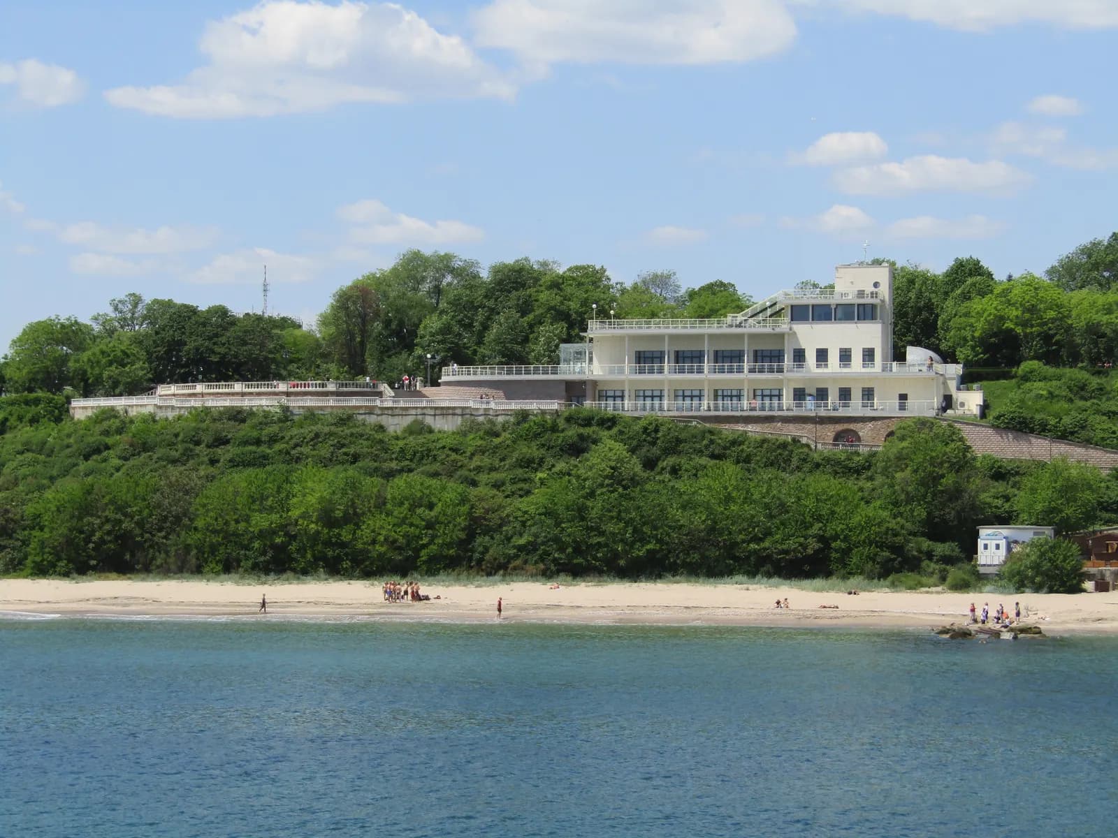 The white Sea Casino building among the trees of the Sea Garden in Burgas, above a sandy city beach, seen across the water from the pier