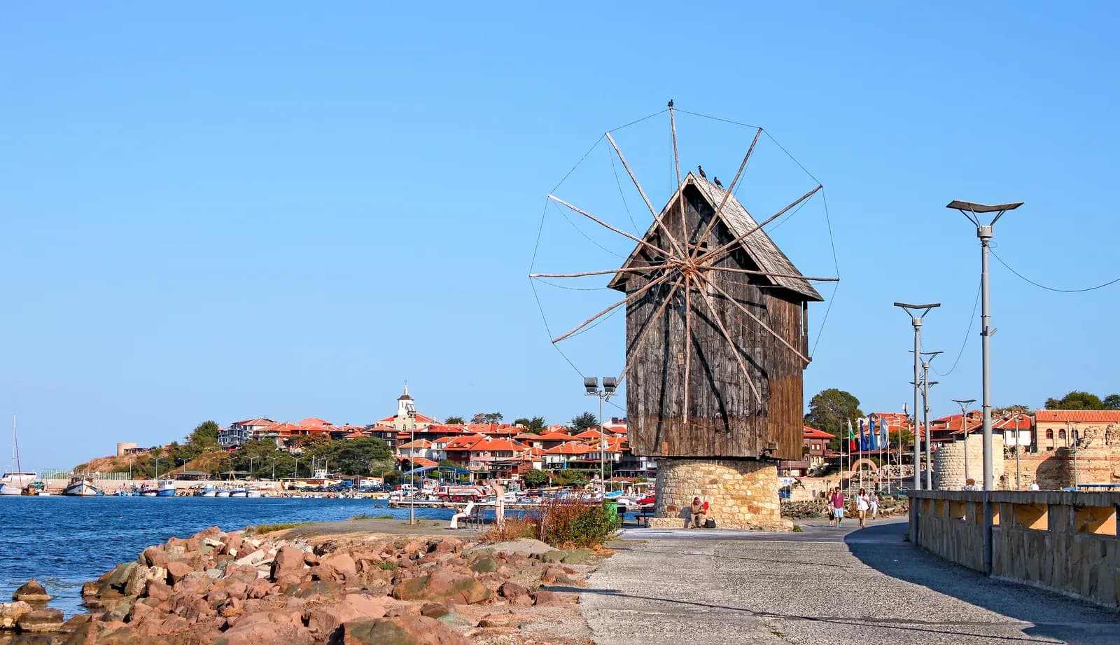 The wooden windmill on the isthmus leading to old Nessebar, with the red roofs and a church bell tower of the old town behind, the harbour on the left and rocks and a stone promenade in front