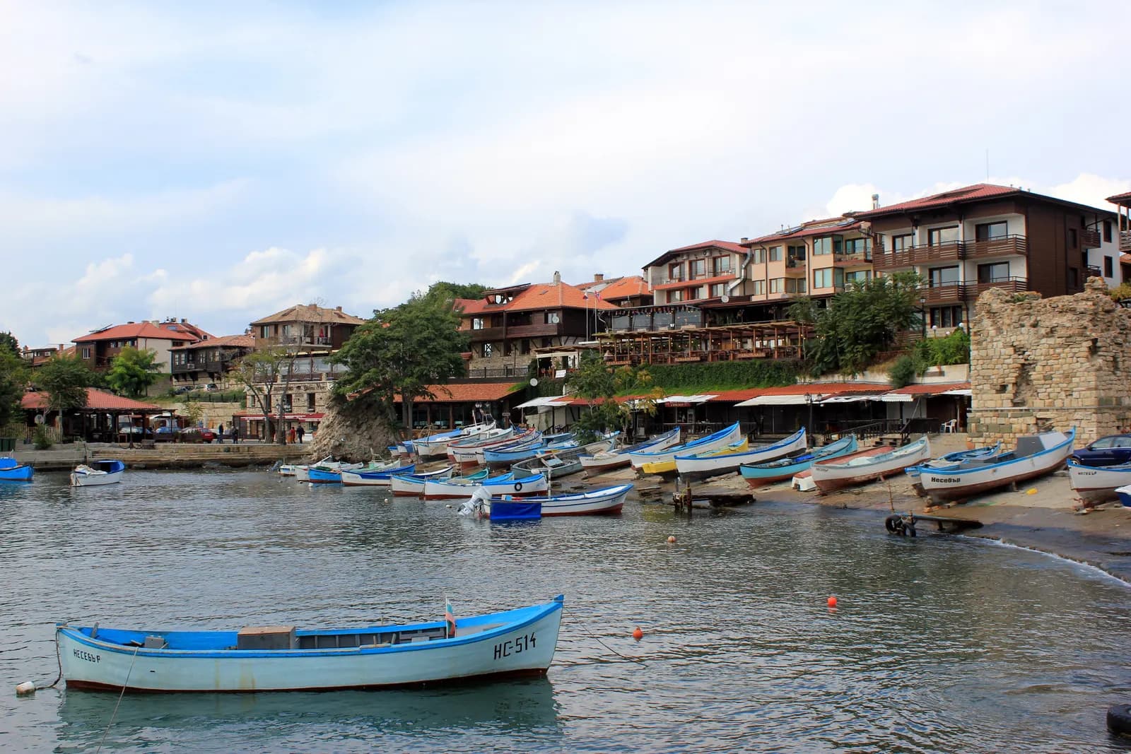 Blue and white wooden fishing boats in the harbour of Nessebar below the stone and timber houses of the old town and a fragment of the old fortress wall