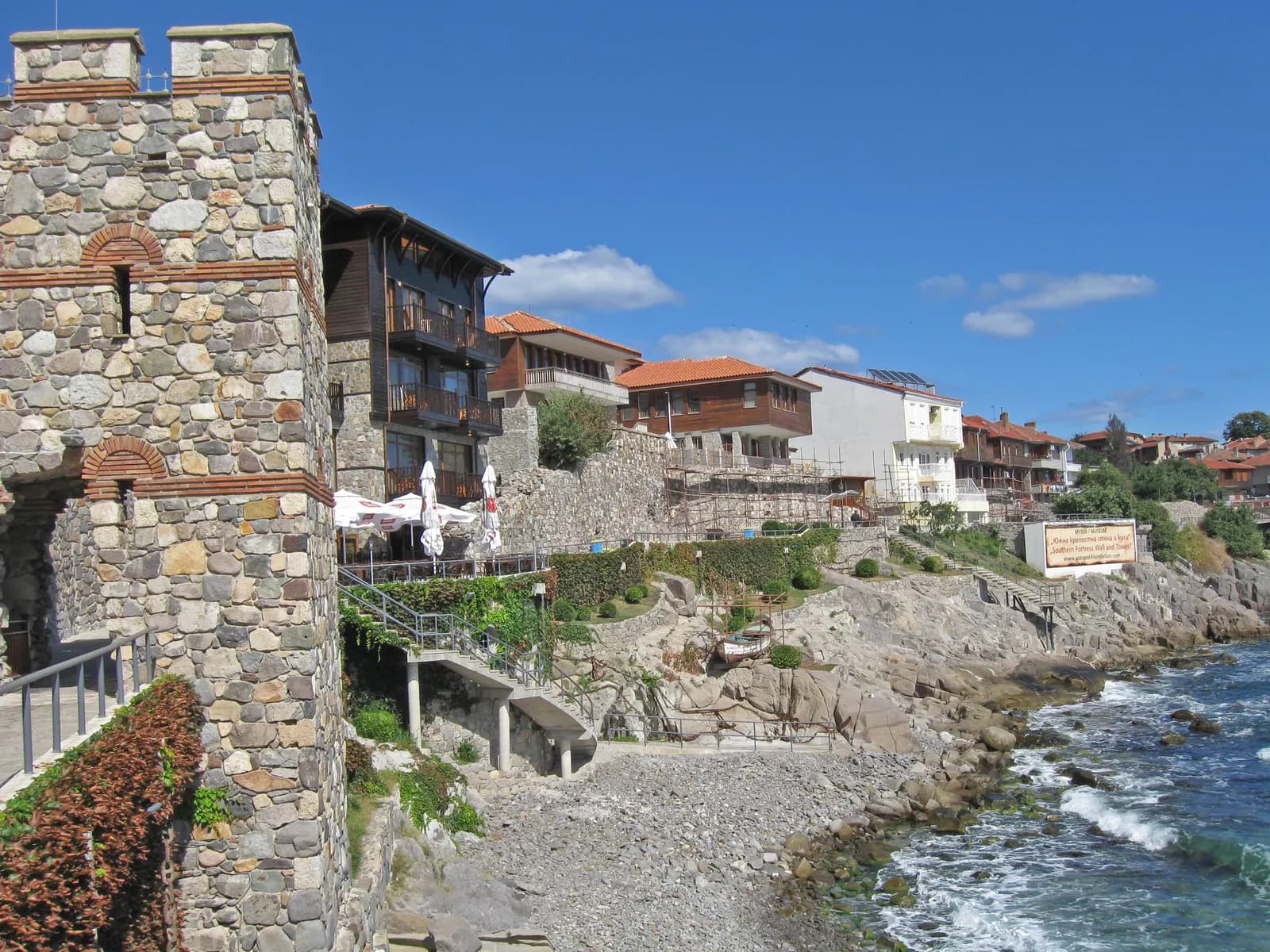 The stone tower of the southern fortress wall in Sozopol beside wooden houses that line the rocky shore, with waves washing over the rocks under a blue sky