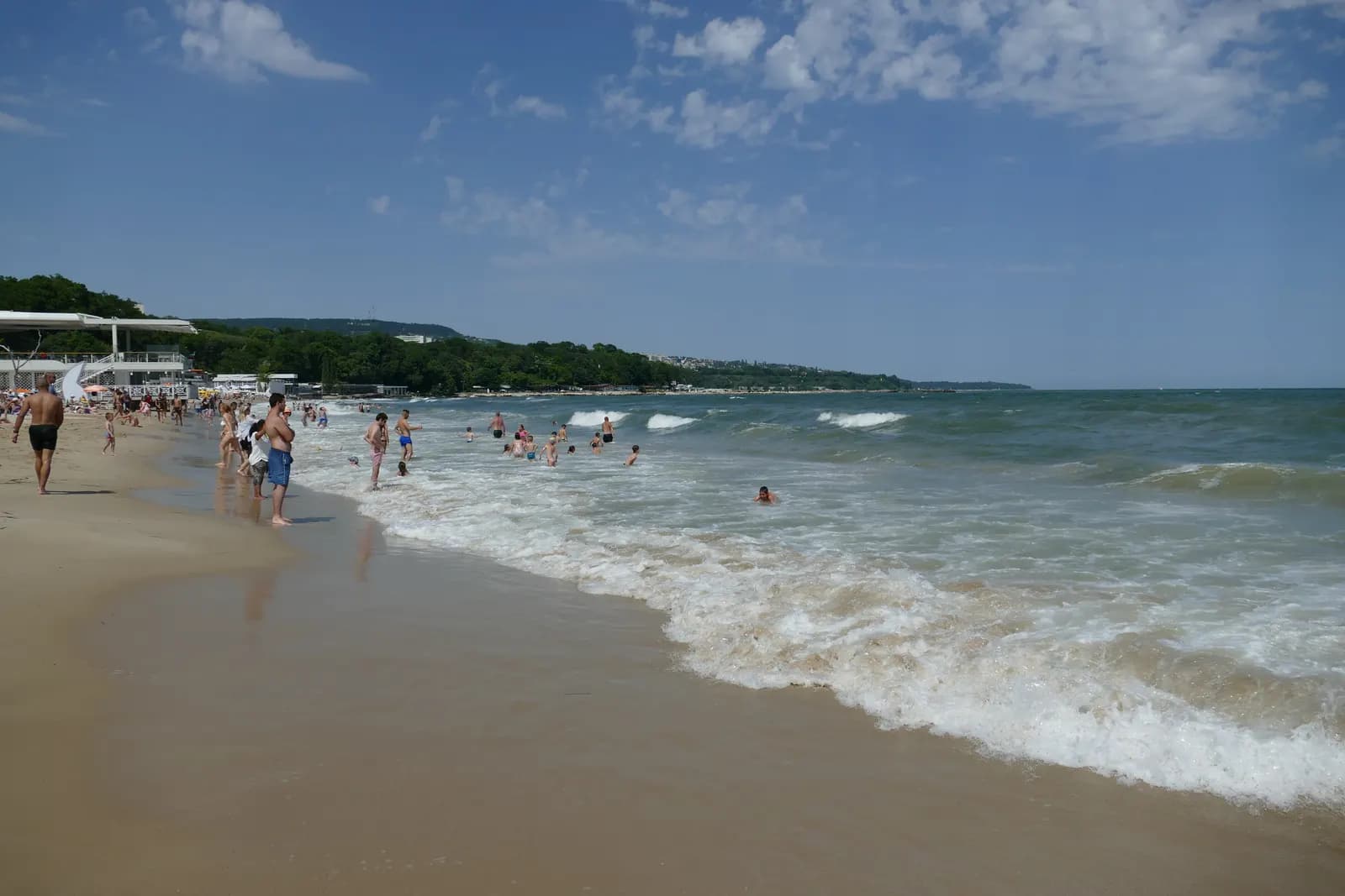 Waves breaking on the sandy city beach below the Sea Garden in Varna, with swimmers in the water and a wooded coastline stretching north under a blue sky