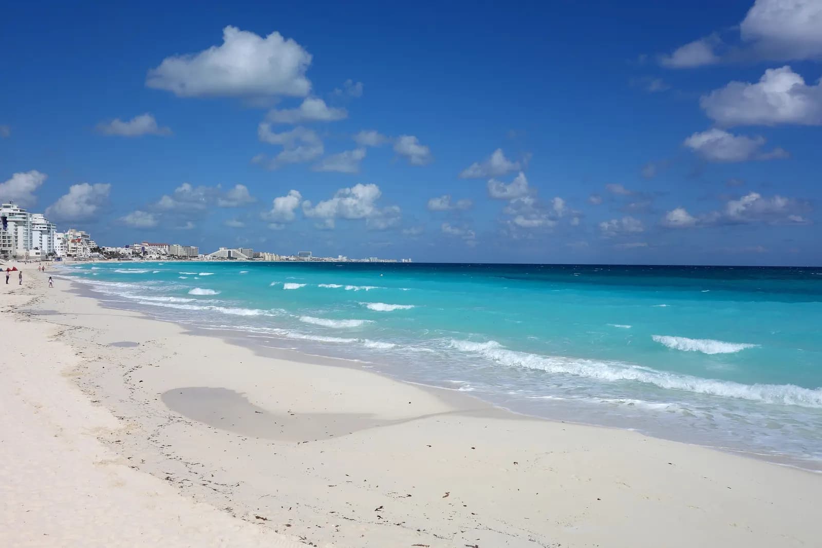 A wide white-sand beach in Cancún with turquoise surf and hotel towers along the shore in the distance
