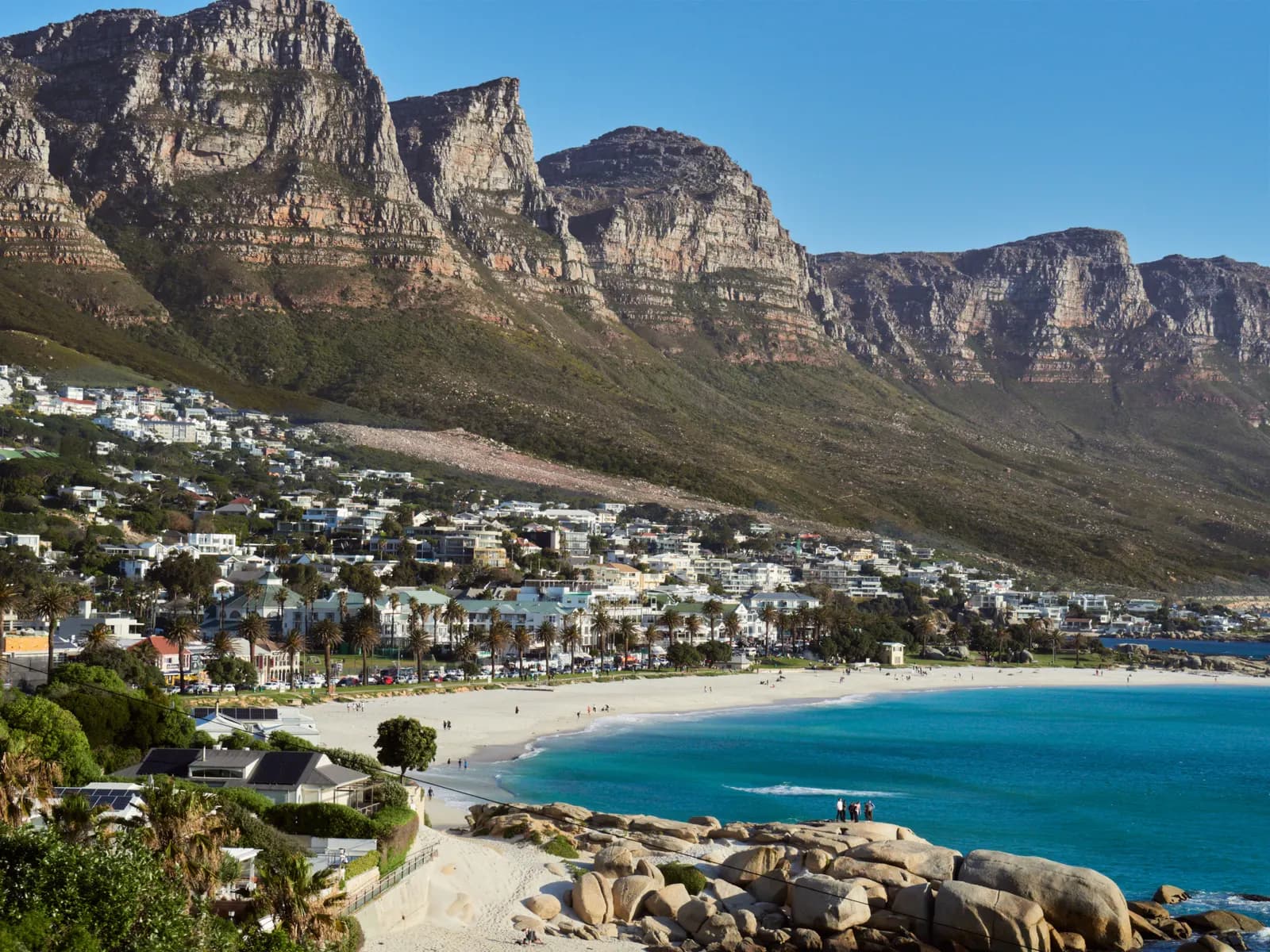 Camps Bay beach and its turquoise bay with granite boulders in front, the houses of the suburb behind and the cliffs of the Twelve Apostles above
