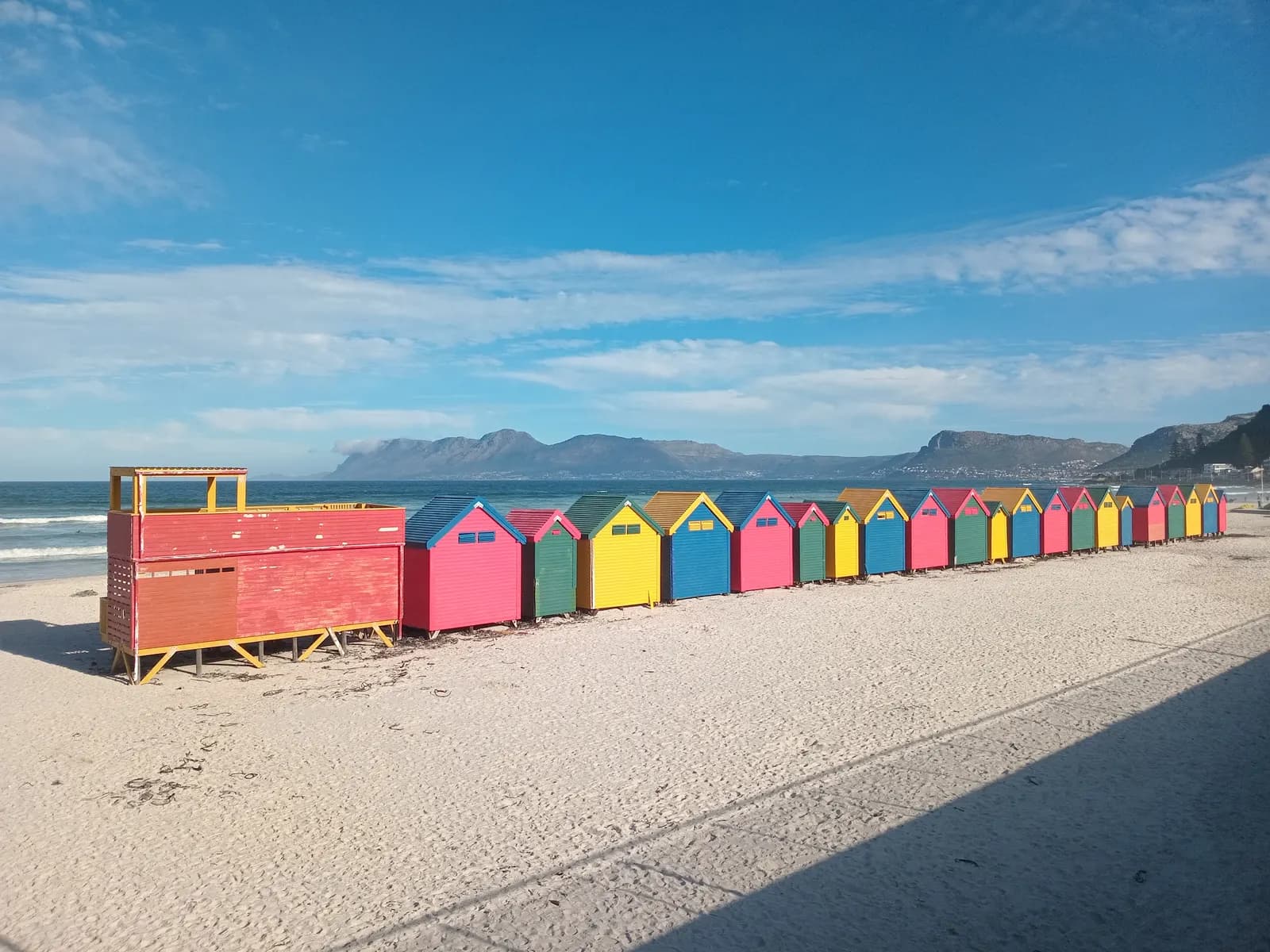 A row of brightly painted beach huts on the sand at Muizenberg, with the sea and mountains behind