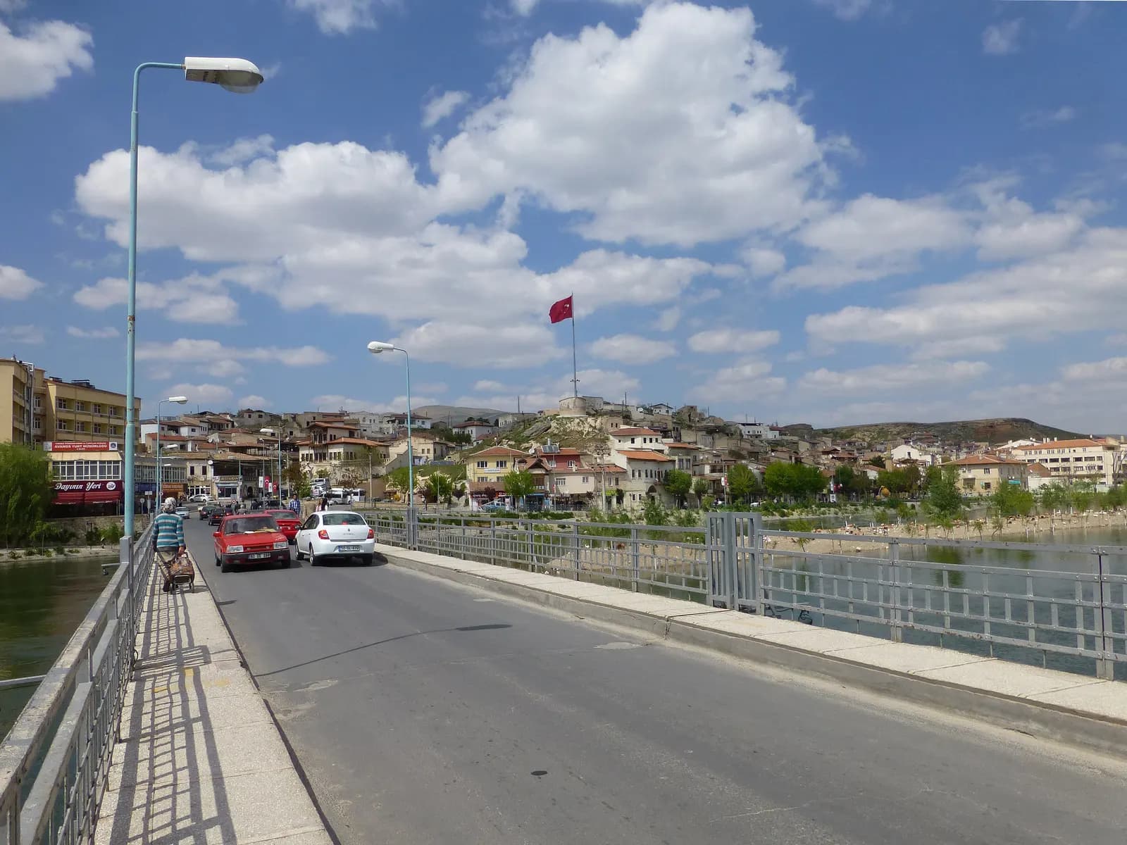 The bridge over the Kızılırmak river into Avanos, with cars crossing and the town climbing the hill behind under a Turkish flag