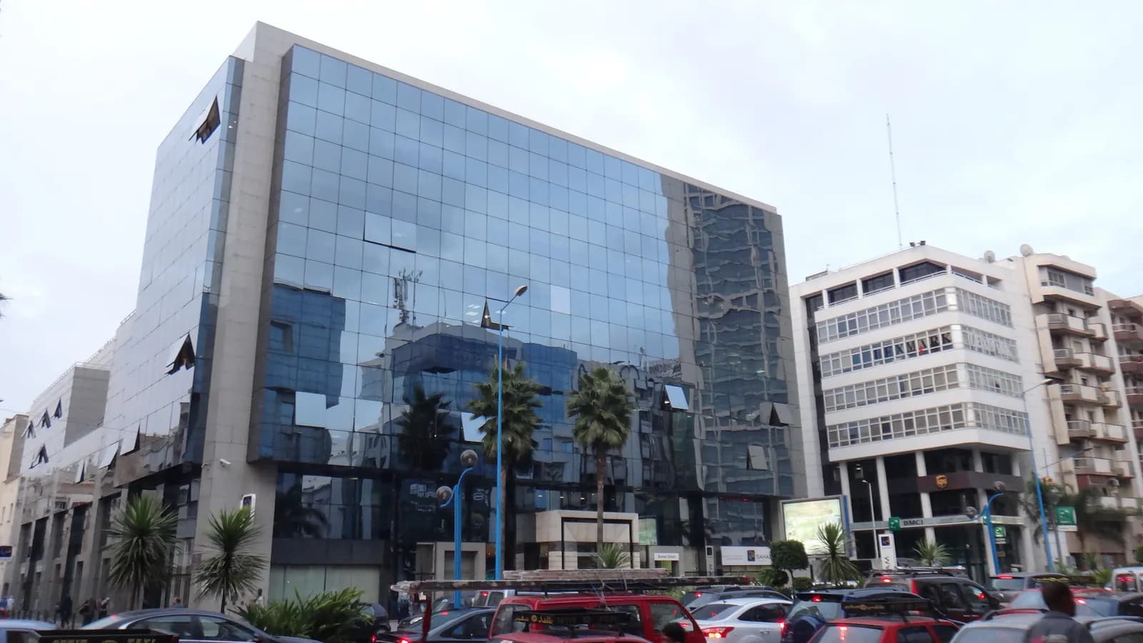 A glass-fronted office building among palms on Boulevard Mohamed Zerktouni in Maârif, with white apartment blocks beside it and traffic in front