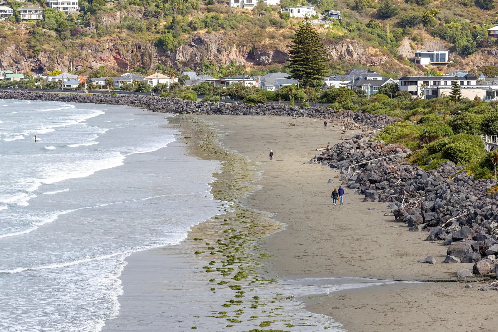 Sumner Beach with surf, a few people walking on the sand, a rock wall and houses on the cliffs above
