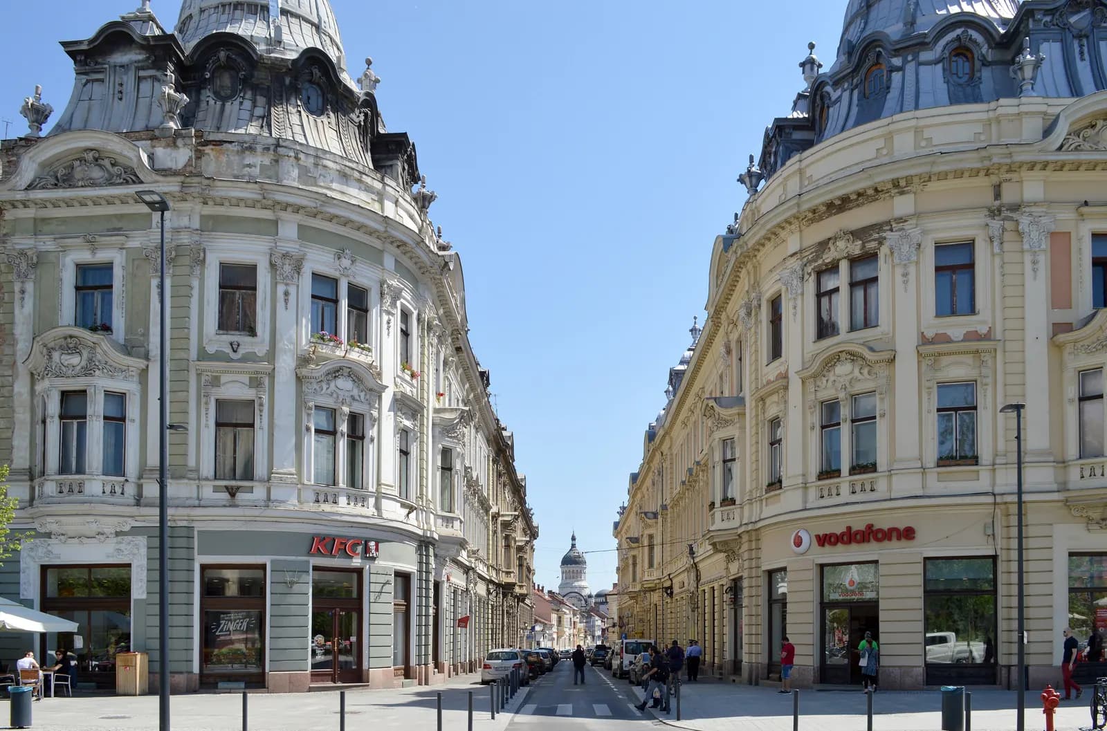 Strada Iuliu Maniu seen from Piața Unirii in Cluj-Napoca, a straight street between two ornate domed corner buildings, with a church dome at the far end