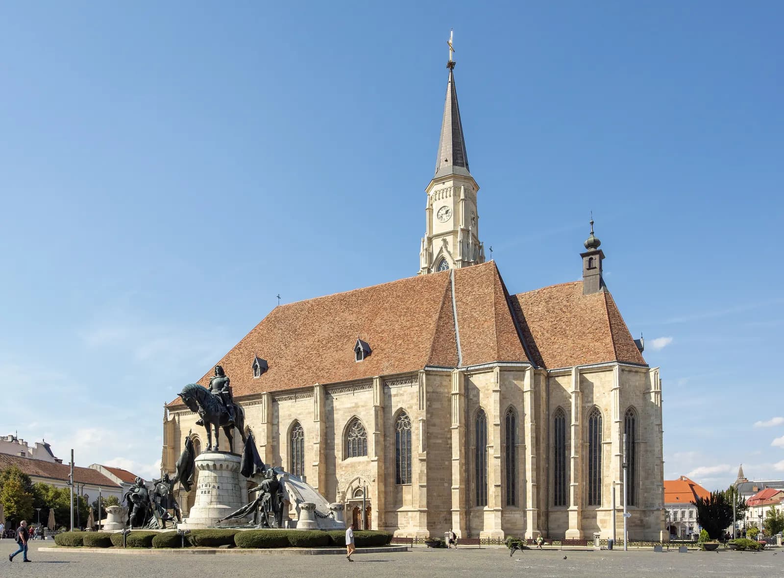 St Michael's Church on Piața Unirii in Cluj-Napoca, a Gothic stone church with a tall spire and a steep tiled roof, with the equestrian Matthias Corvinus monument in front