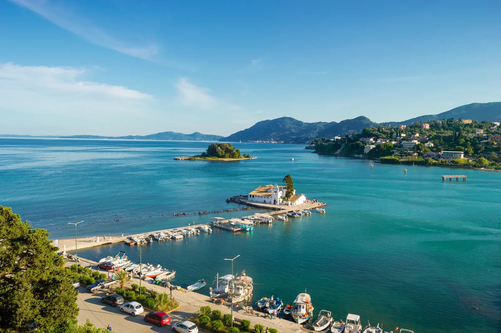 The Vlacherna monastery at the end of its causeway at Kanoni, with Pontikonisi islet behind, boats moored along the quay and wooded hills across the bay