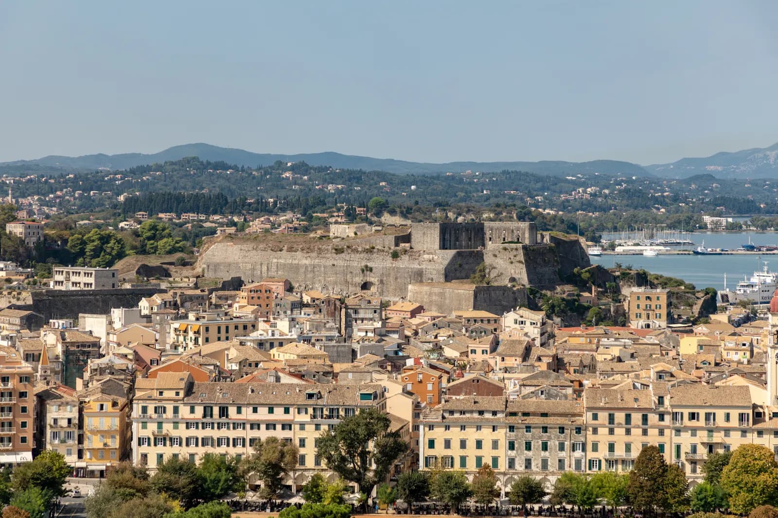 Tiled roofs and shuttered facades of Corfu old town, with the walls of the New Fortress, the harbour and green hills behind