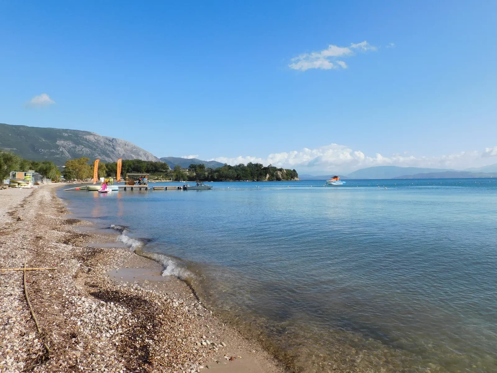The shingle beach at Dassia on the east coast of Corfu, with a wooden jetty, a wooded headland and the mountains of the mainland across calm water