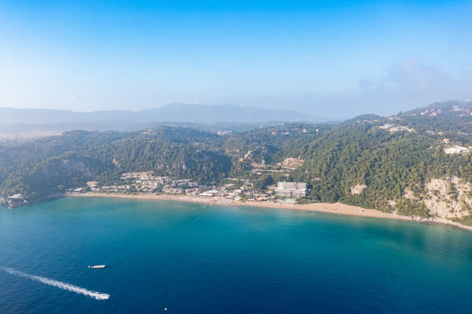 Aerial view of Glyfada beach on the west coast of Corfu, a long strip of sand below wooded hills with hotels and apartments behind it