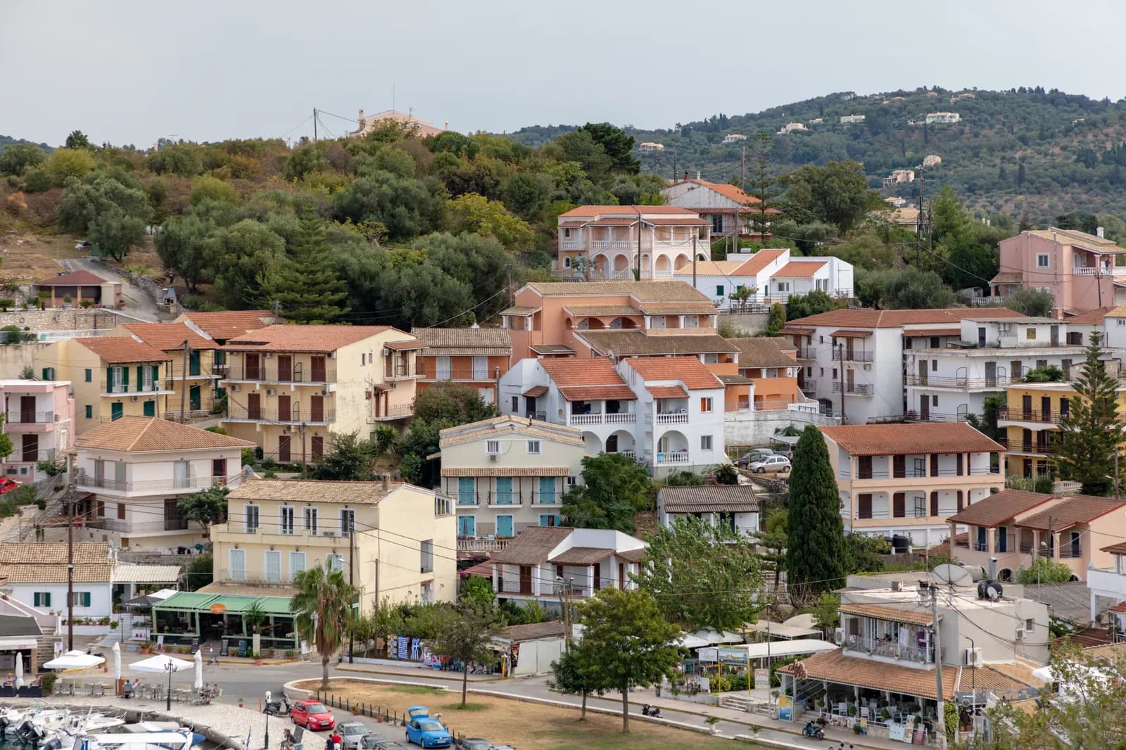 Houses and apartment buildings with tiled roofs on the hillside above the harbour front in Kassiopi, with olive groves on the slopes behind