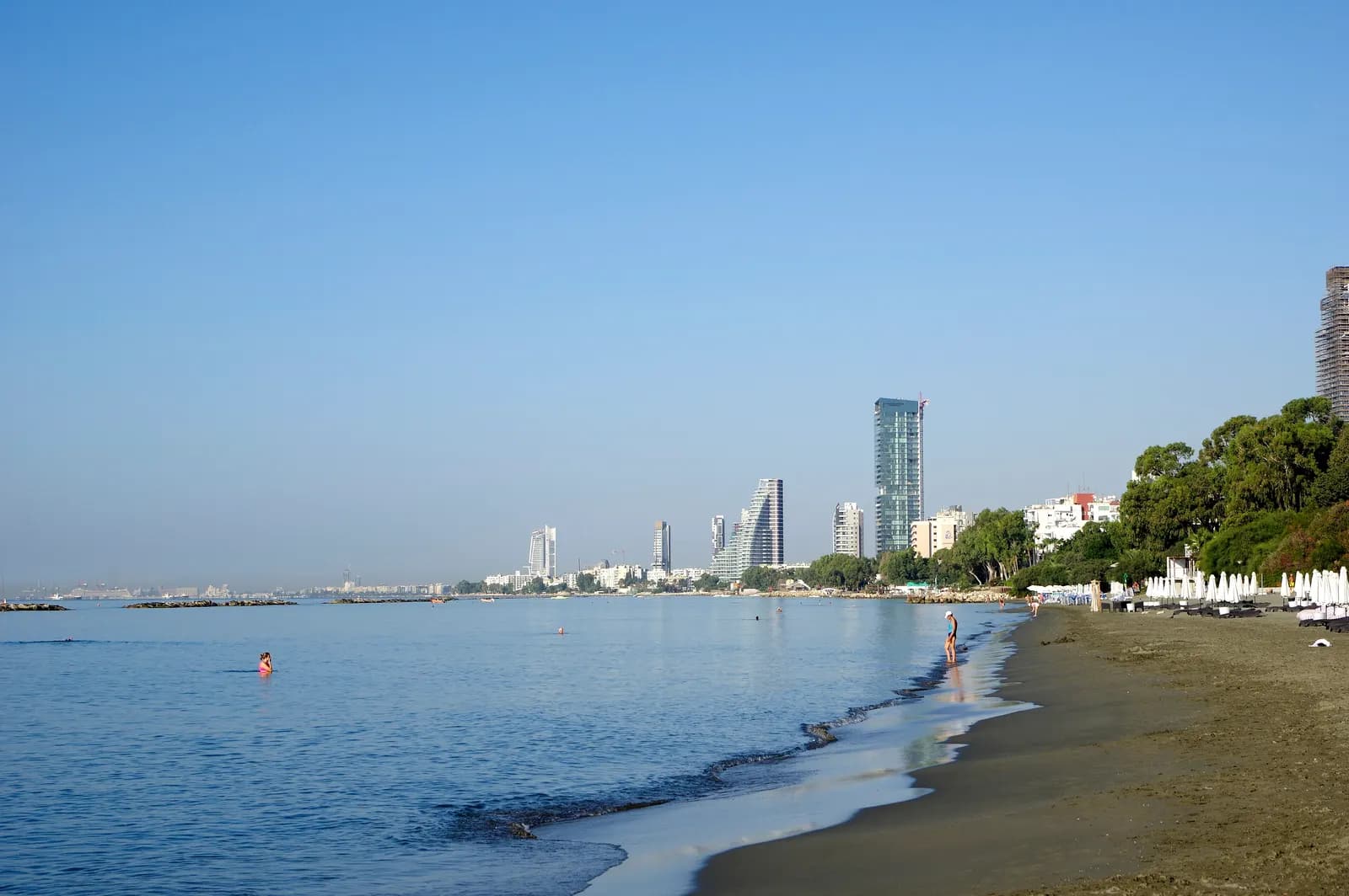 A dark sand beach in Limassol in the morning, with calm water, a few swimmers and the high-rise towers of the seafront in the distance