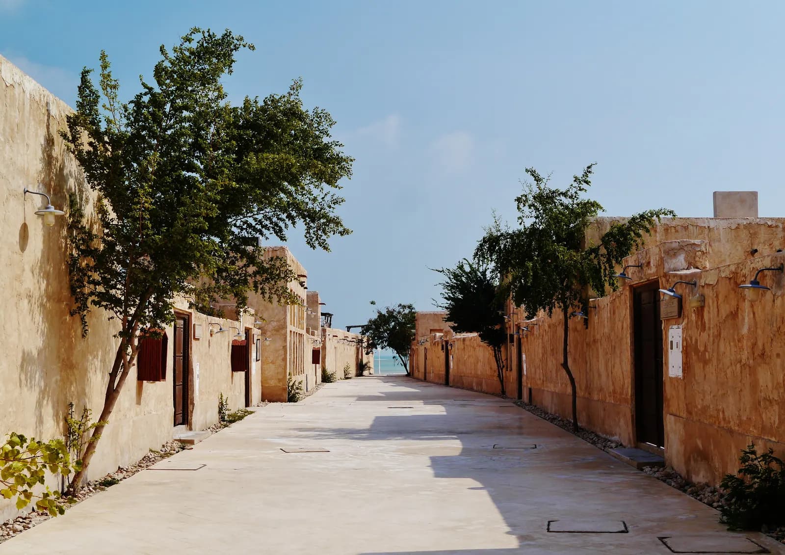 A paved lane between sand-coloured walls and trees in the old souq of Al Wakrah, with the sea at the end of the lane
